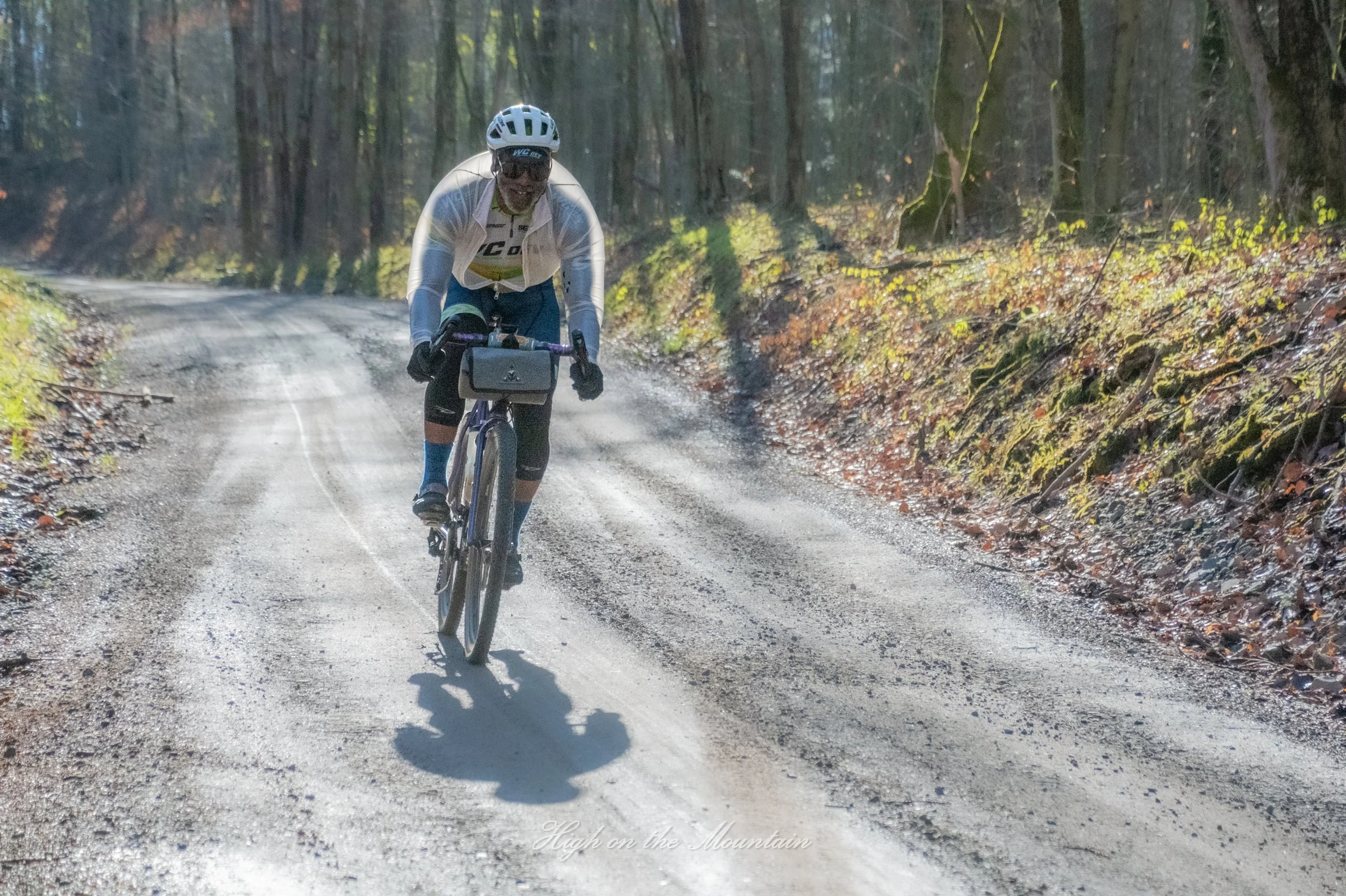 A person riding a mountain bike on a dirt trail in a forest during autumn. They are wearing a helmet, sunglasses, and cycling gear, smiling and focused on riding.