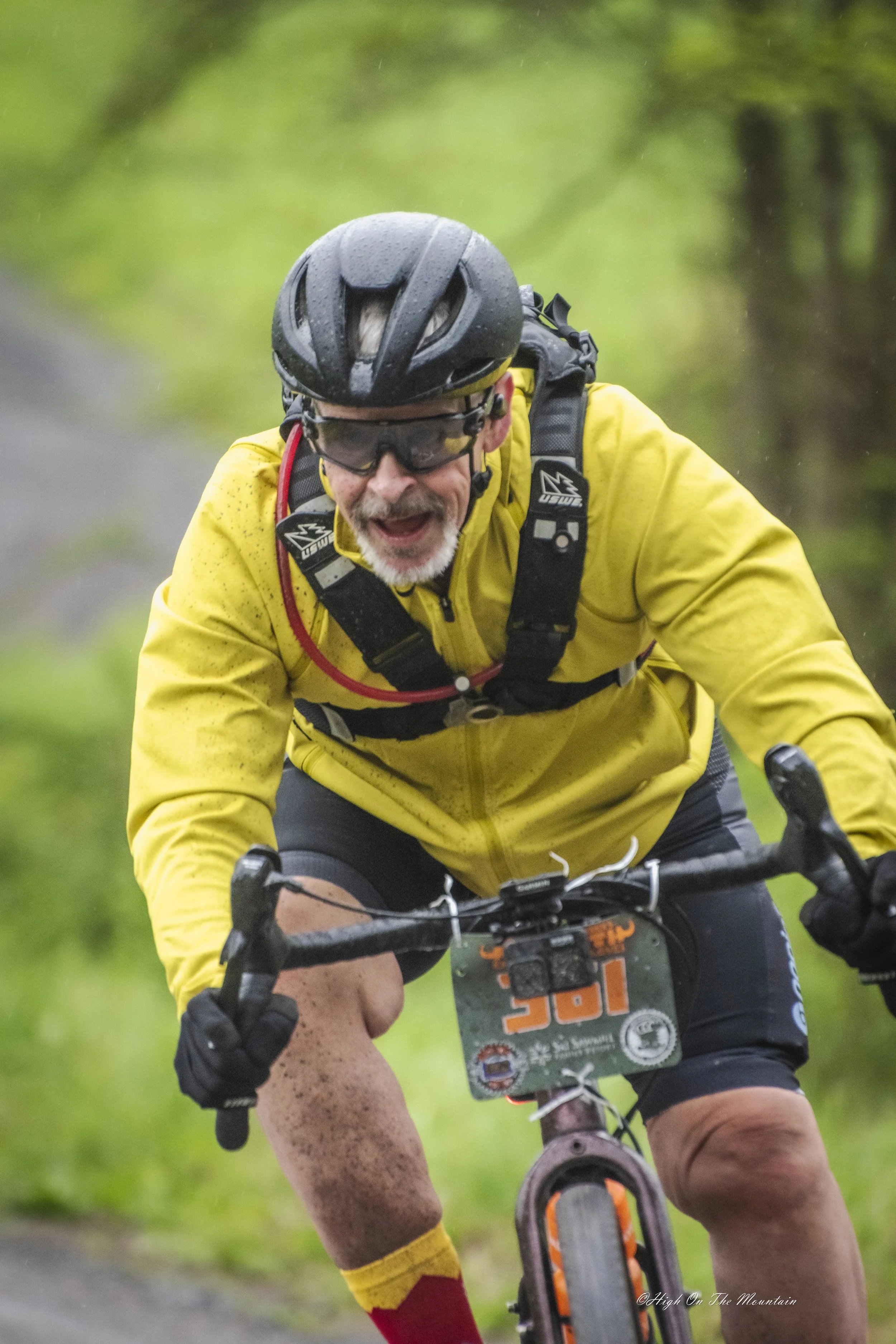 An older man wearing a yellow jacket, black helmet, and sunglasses rides a mountain bike on a trail in a green forested area, smiling and enjoying the ride.