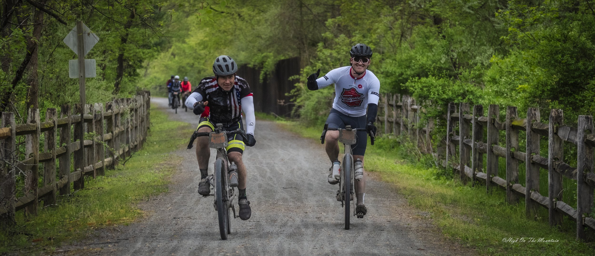 Two cyclists, wearing helmets and cycling gear, ride on a gravel trail surrounded by green trees. Other cyclists are visible in the background.