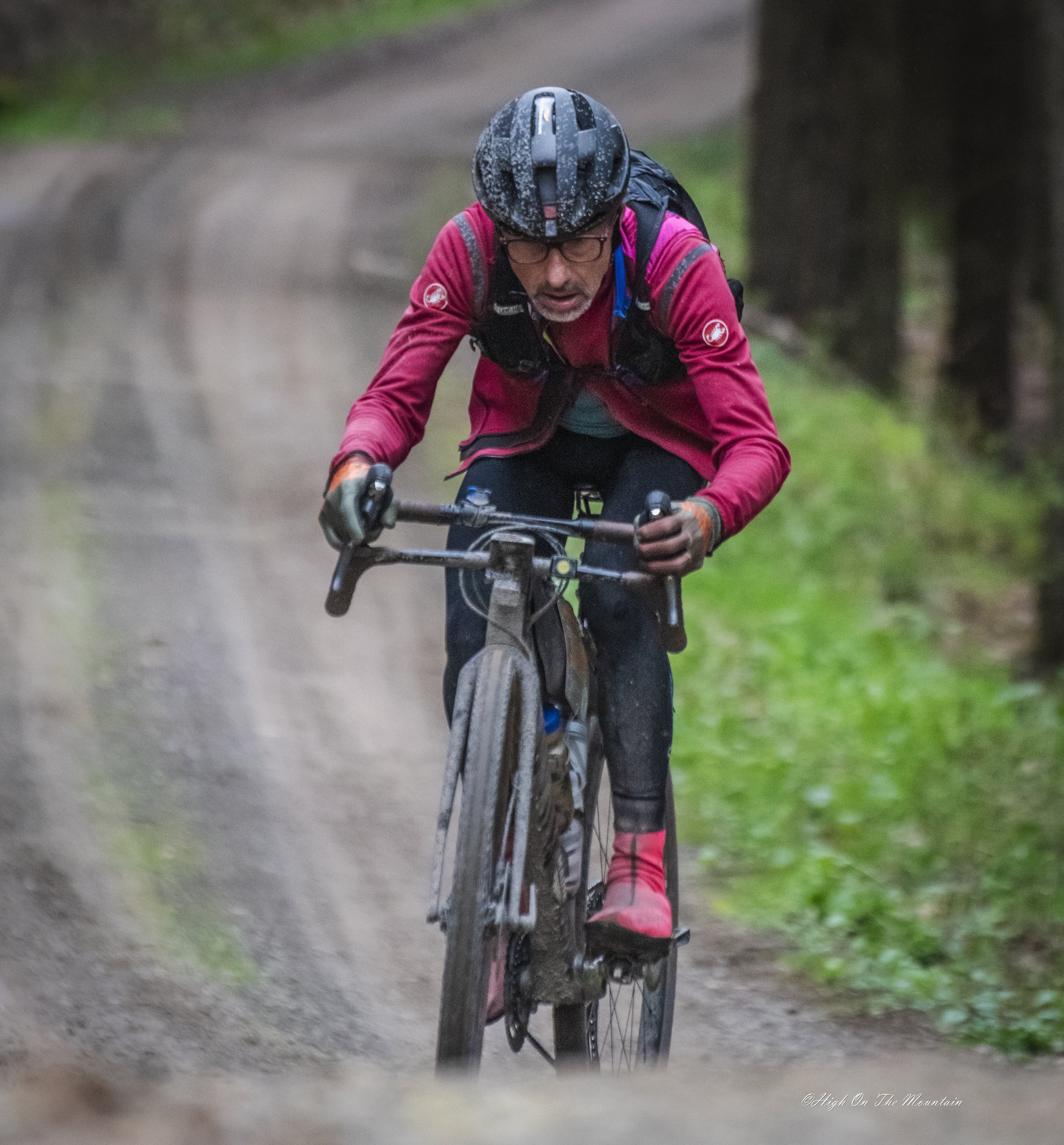 A man riding a mountain bike on a muddy trail in a forest, wearing a black helmet, glasses, a red jacket, and gloves.