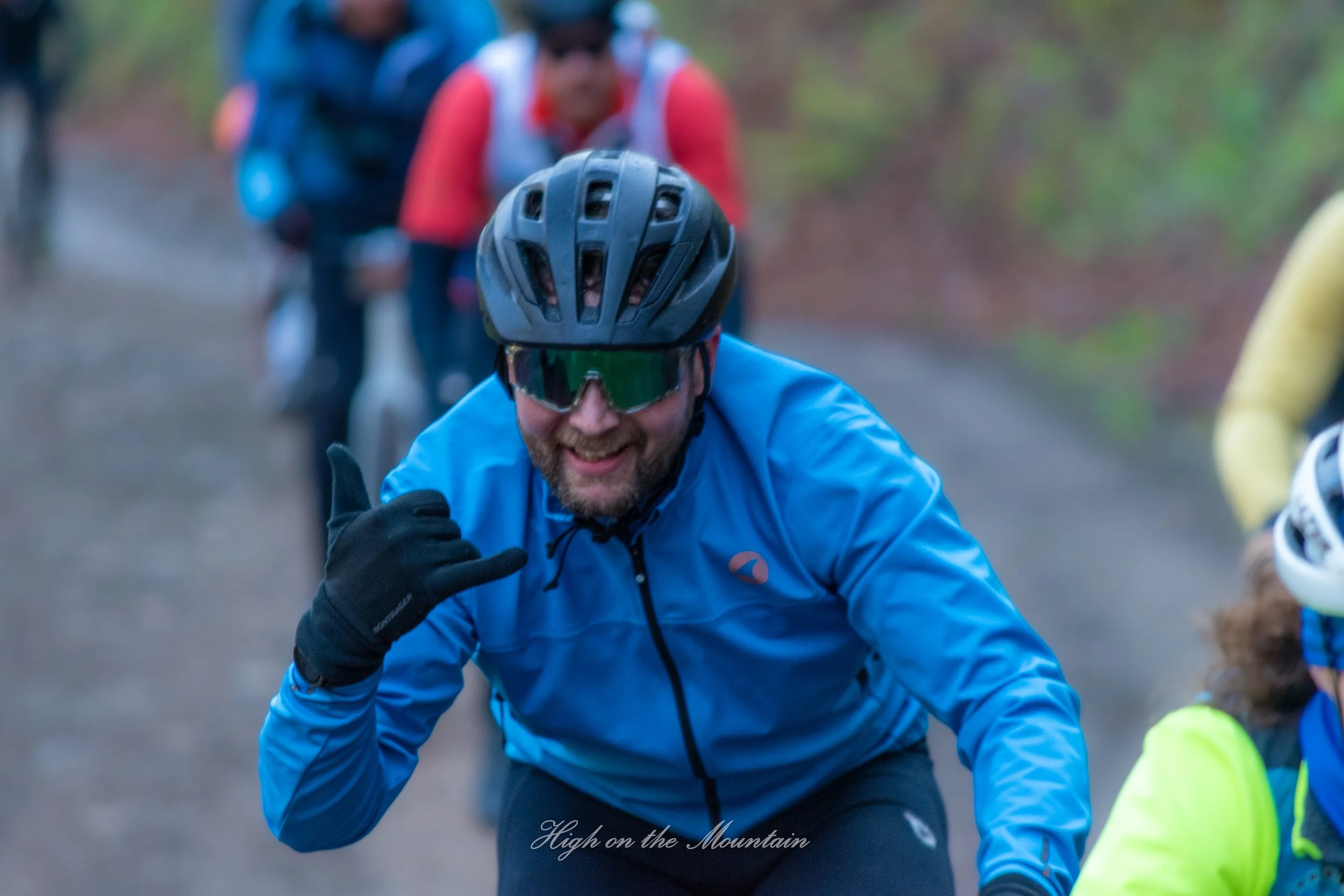 A man wearing a black helmet and sunglasses smiling and giving a thumbs up while riding a bicycle outdoors, with a blurred background of other cyclists and trees.