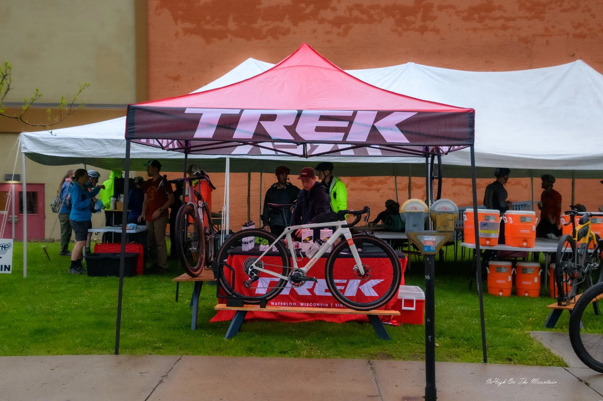 Outdoor booth with two tents, one red with 'TREK' branding and one white, set up on green grass. Several people are gathered around, some wearing helmets. Bicycles and equipment are displayed under the tents, along with water containers. In the backg