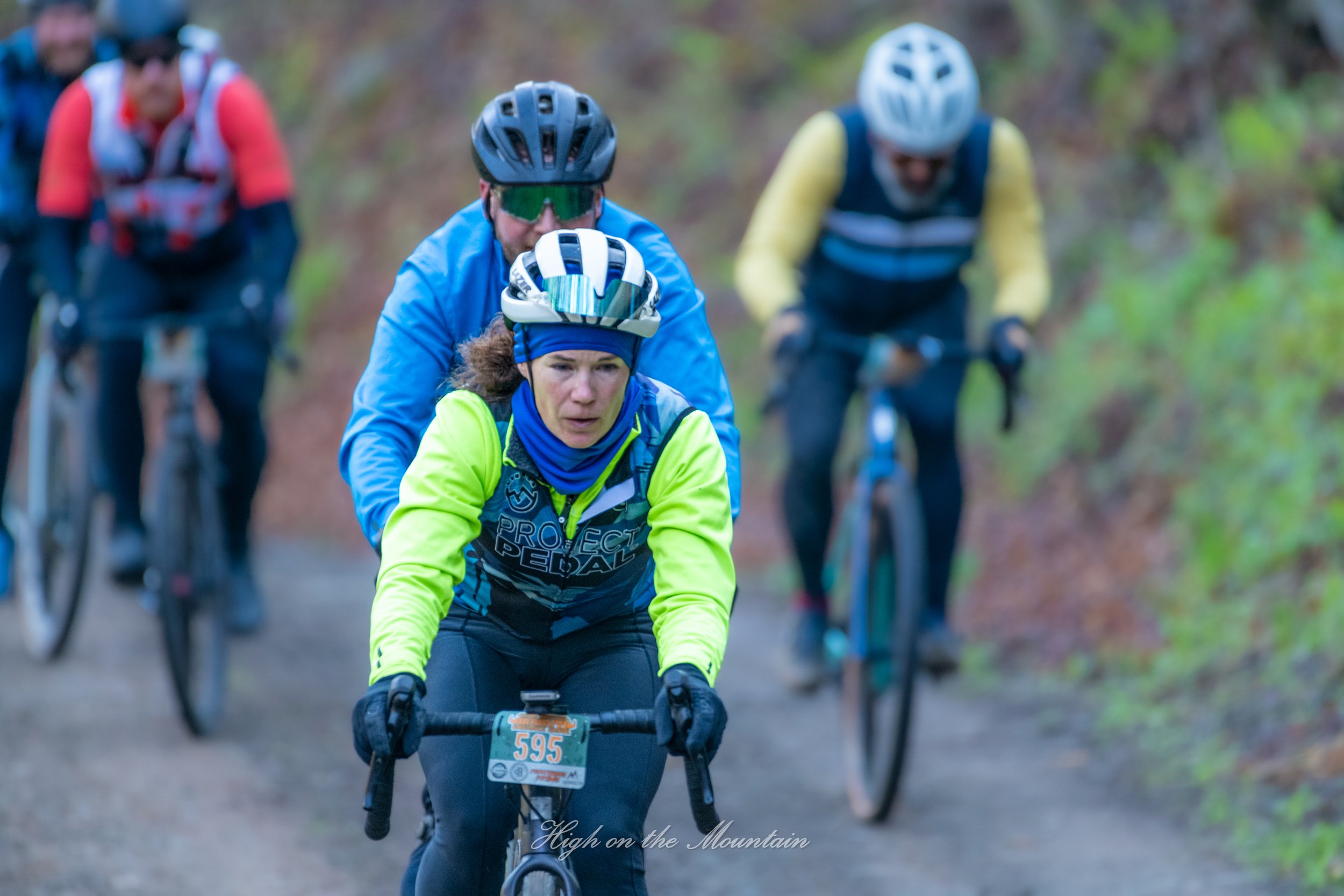 A group of mountain bikers riding on a trail in a forest, with one woman in a blue and yellow outfit and a helmet in the foreground, leading the group.