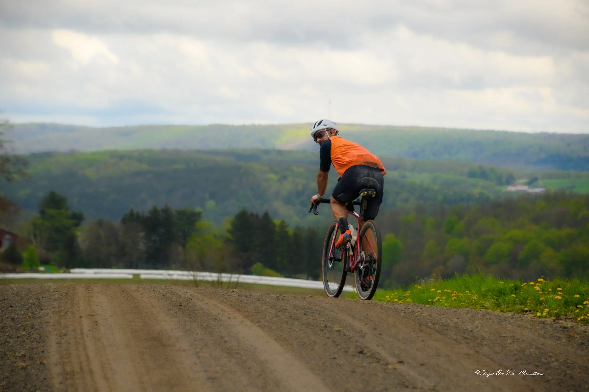 A cyclist wearing an orange and black outfit and a white helmet rides a bicycle on a dirt road with hills and green trees in the background under a cloudy sky.