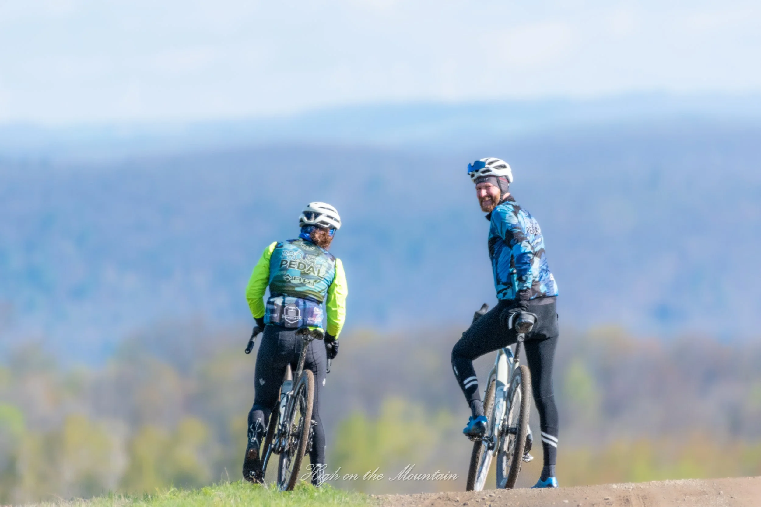 Two people riding bicycles on a mountain trail, wearing helmets and sports gear, smiling as they enjoy a sunny day outdoors.