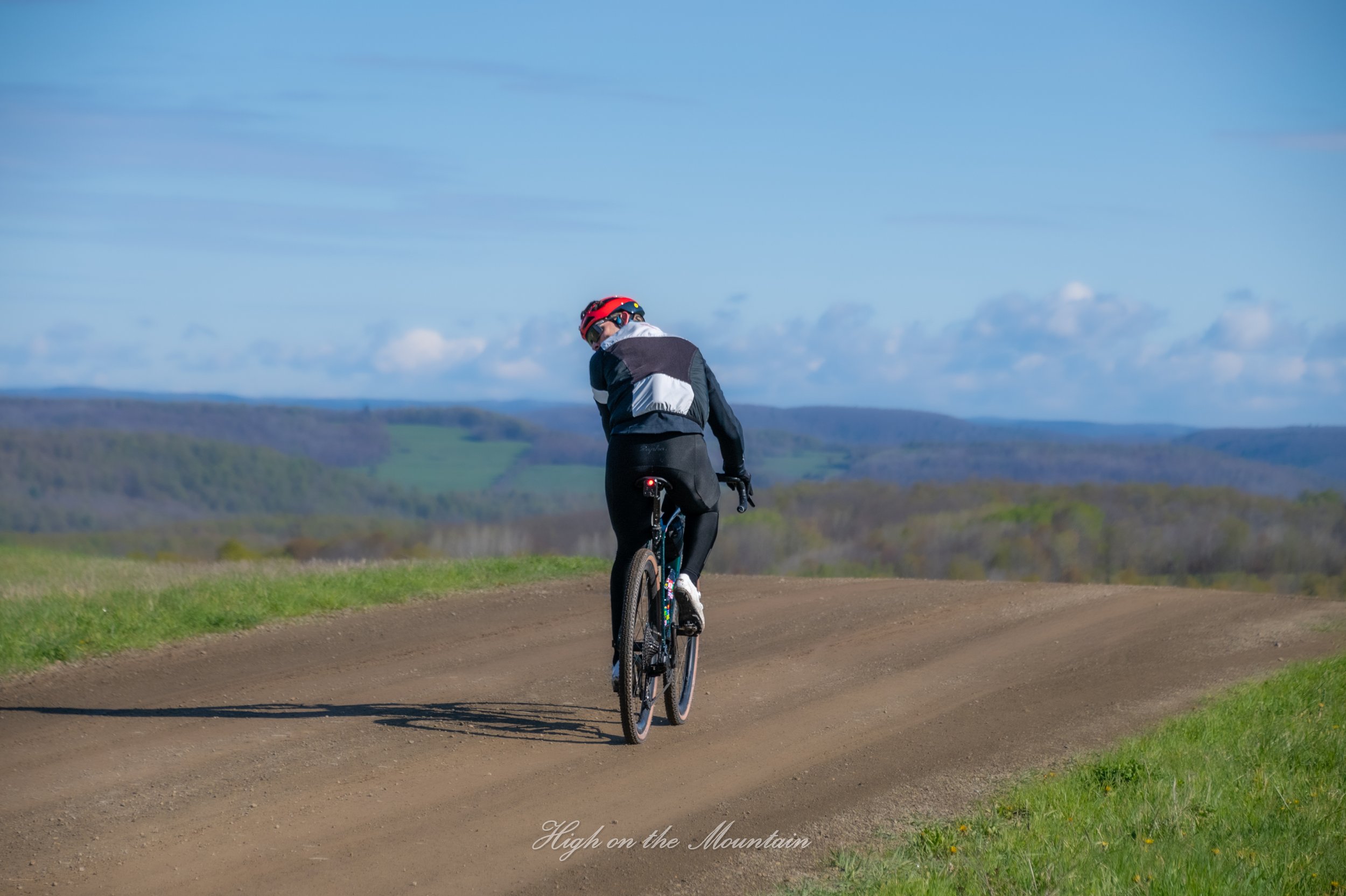 A person riding a bicycle on a dirt path through a landscape of green hills and a blue sky with some clouds.