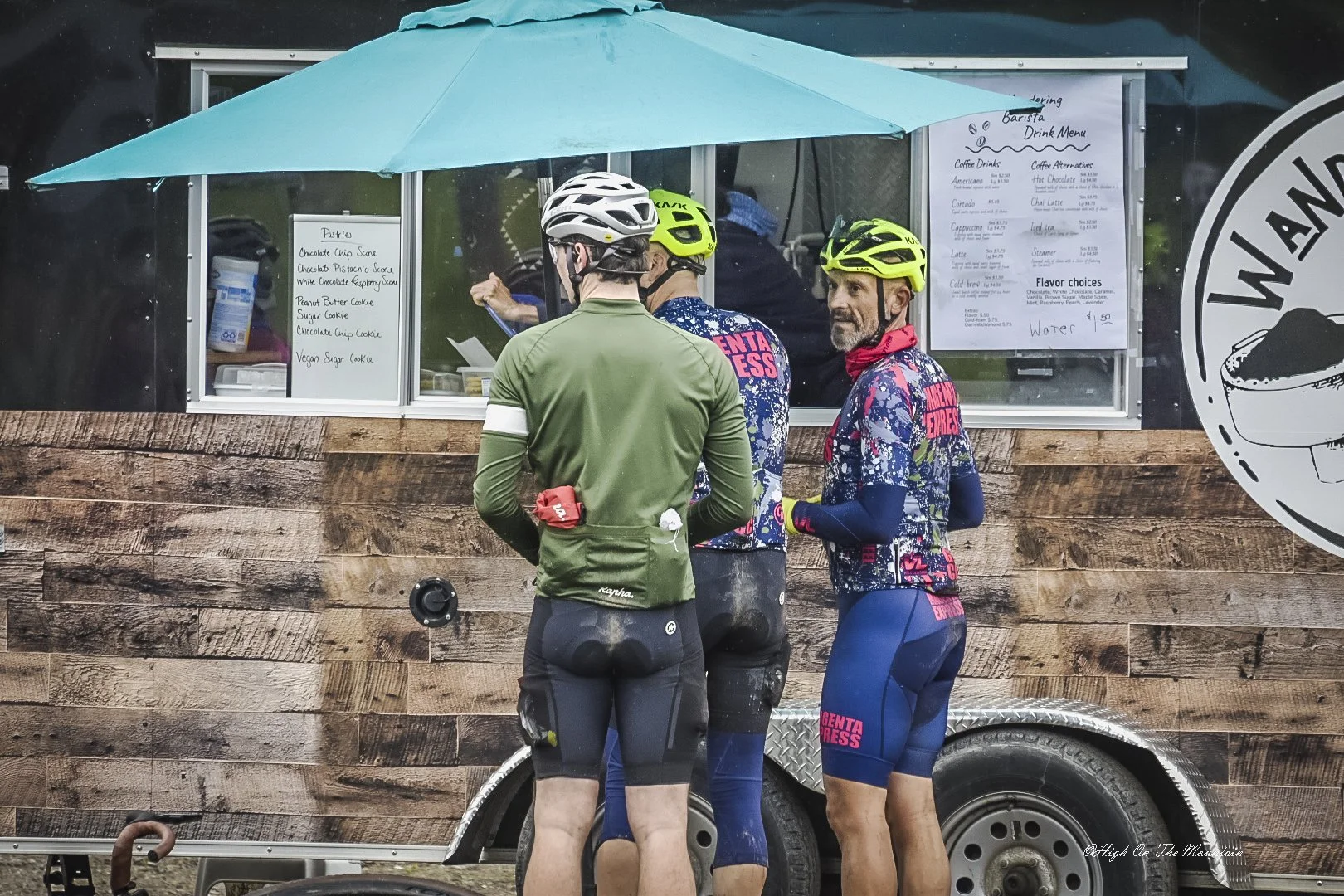 Three cyclists in gear standing in front of a food truck, with a menu visible, under a blue umbrella.