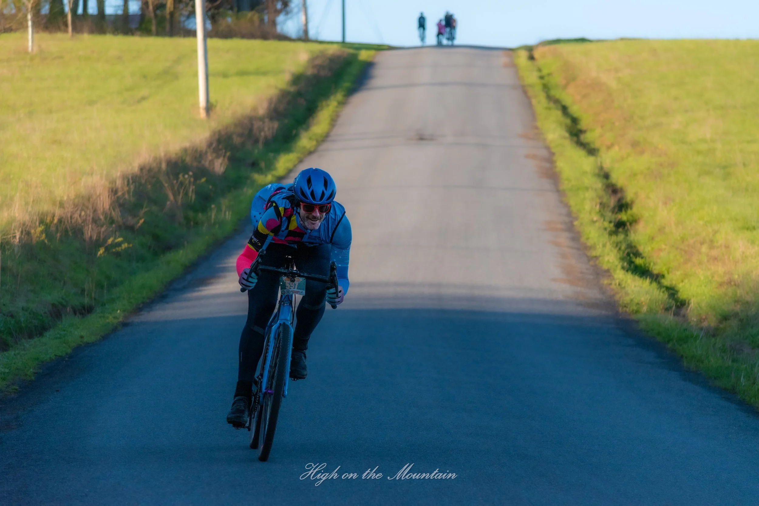 A cyclist wearing a blue helmet, sunglasses, and a colorful jacket riding a bike on a hilly road with grass on both sides during daytime, with a group of people in the background at the top of the hill. Text at the bottom reads "High on the Mountain.