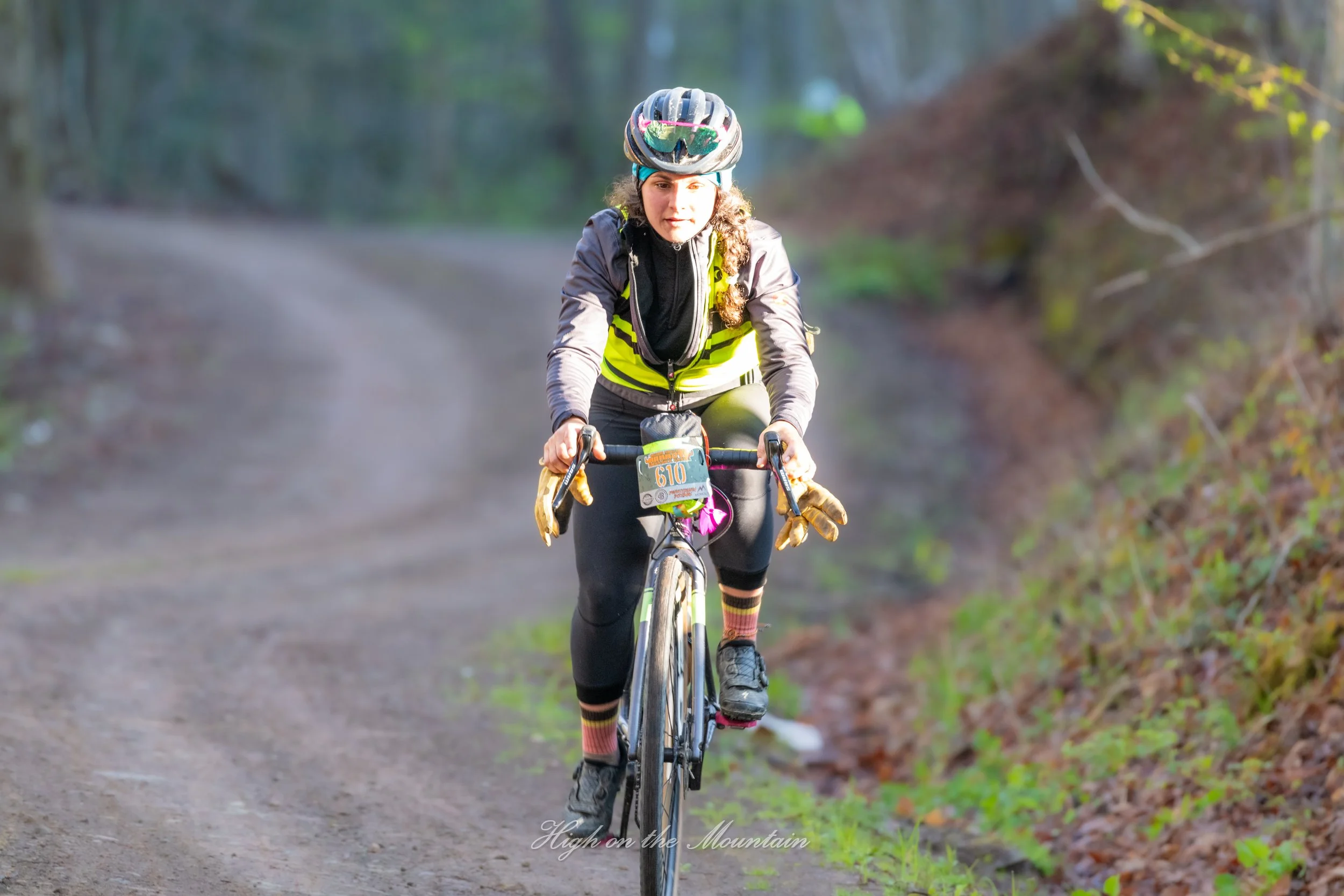 A woman riding a mountain bike on a trail in a wooded area, wearing a helmet, sunglasses, and cycling gear.