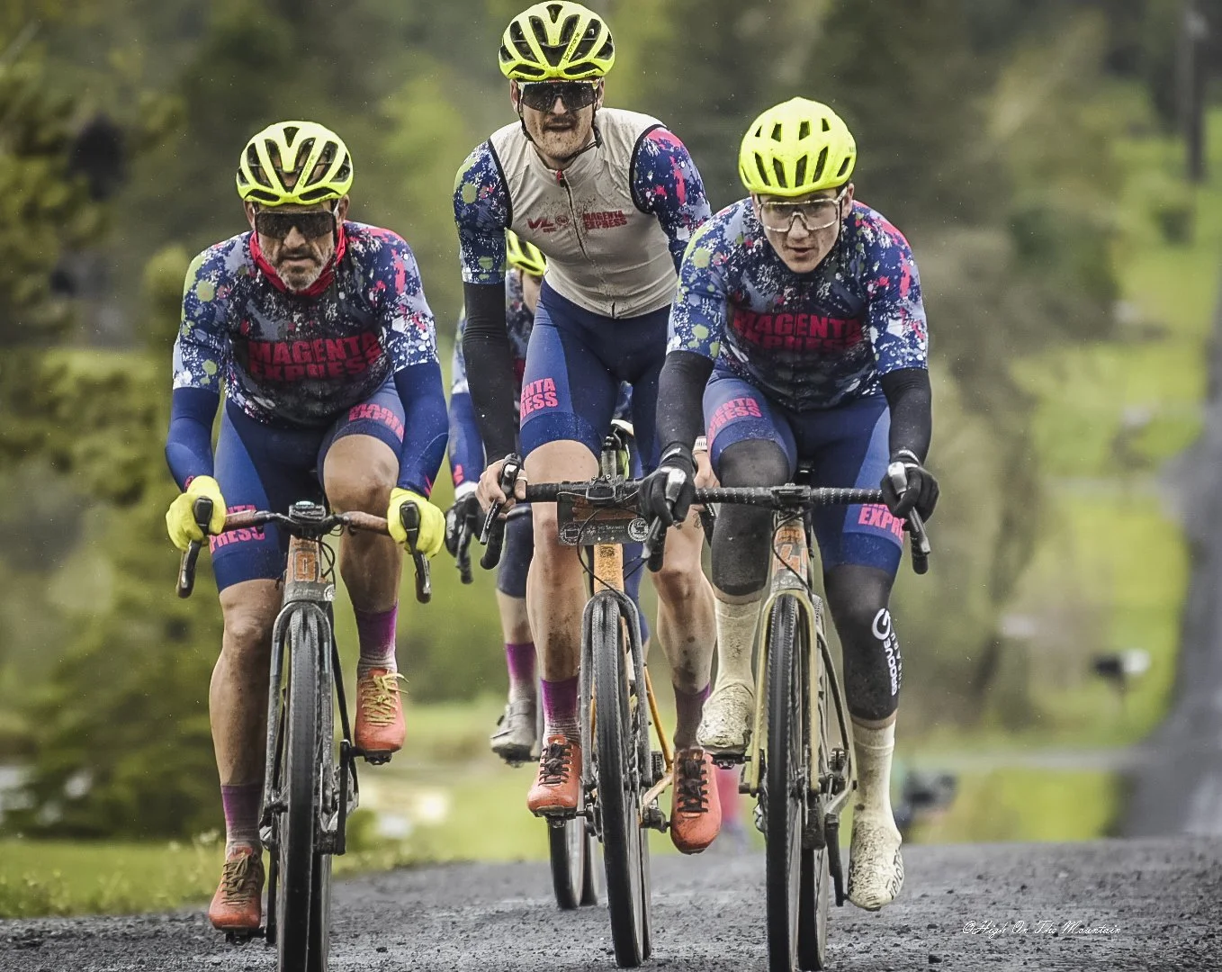 Three cyclists racing on a muddy trail in a forest, wearing bright yellow helmets, sunglasses, and matching blue and pink jerseys.