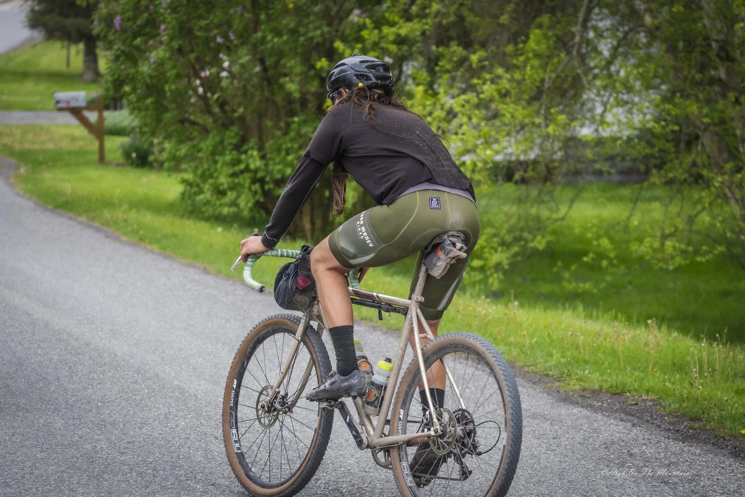 A person riding a mountain bike on a paved trail surrounded by green trees and grass, wearing a black helmet, black jacket, and green shorts.