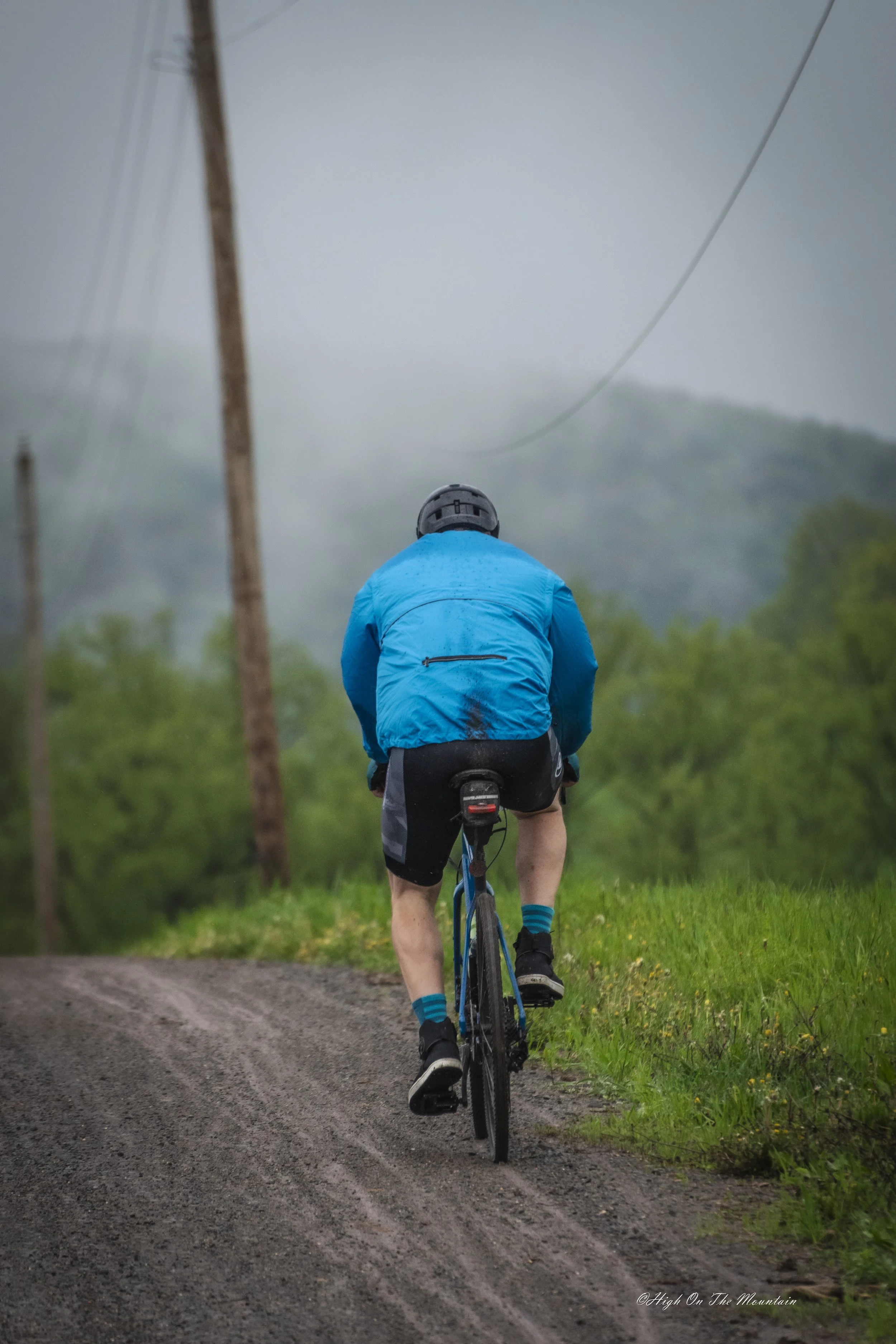 A person wearing a blue jacket, black shorts, and a helmet riding a mountain bike on a dirt trail through green fields with trees and foggy mountains in the background.
