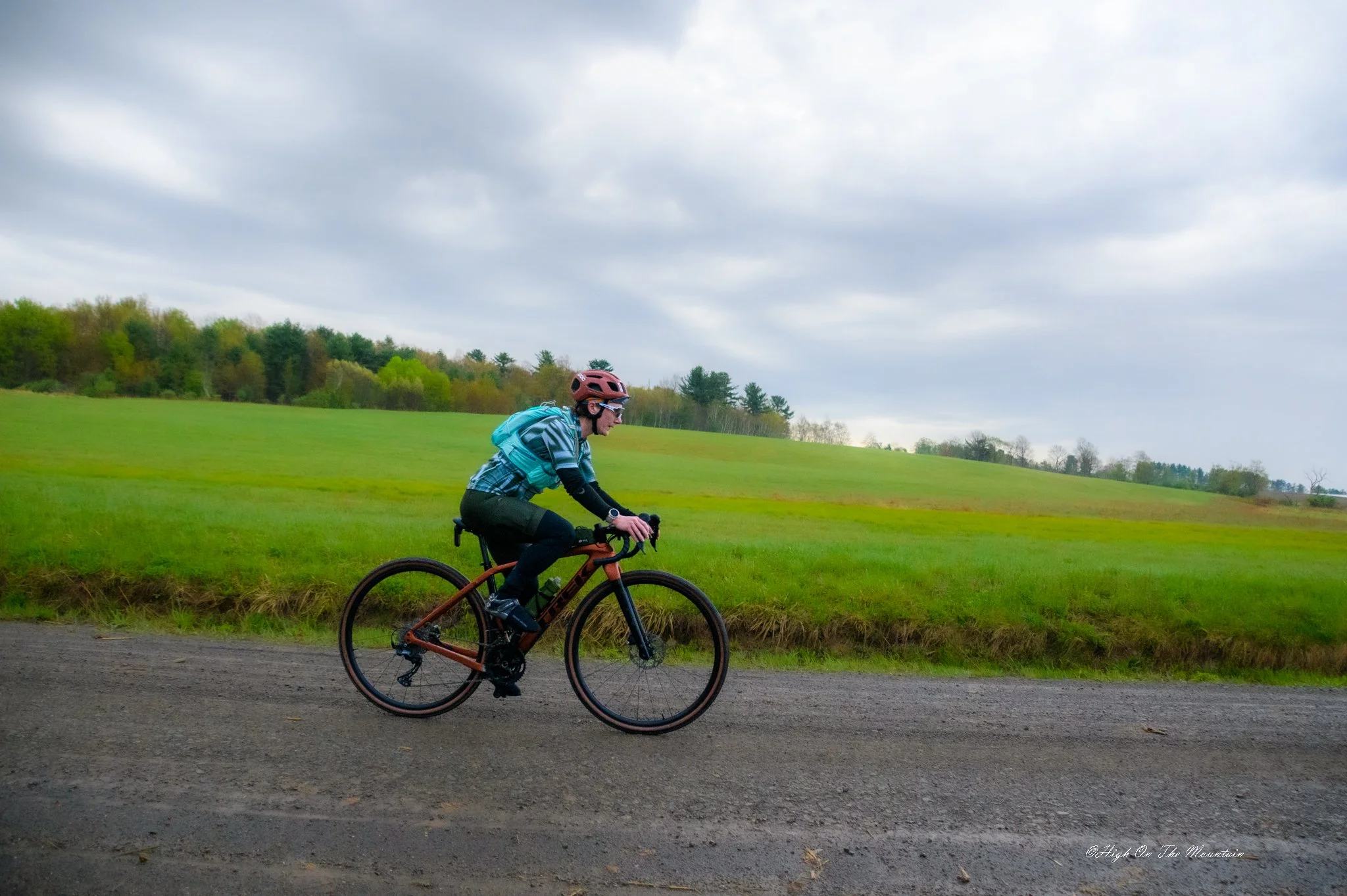 A person riding a bicycle on a paved path through a green, open field with trees in the background and a cloudy sky overhead.