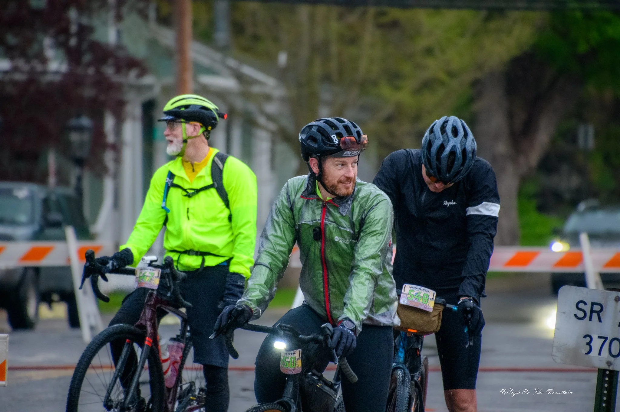 Three male cyclists wearing helmets and cycling gear standing with their bikes behind a barrier, possibly at the start or end of a race, with trees and buildings in the background.