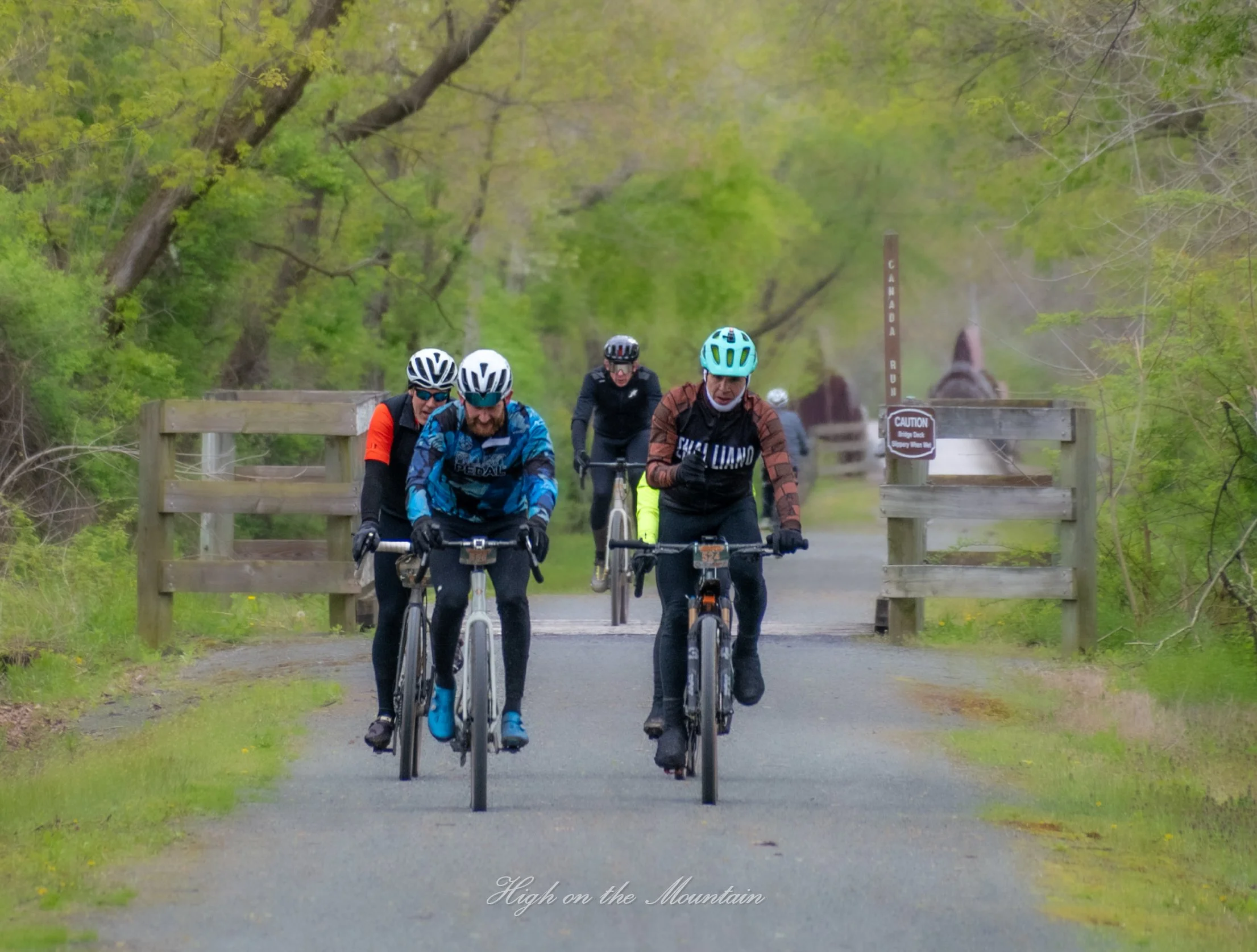 A group of five cyclists riding their bikes on a paved trail through a green wooded area, with a wooden railing and a sign that reads "Canaborah" and a caution sign visible in the background.