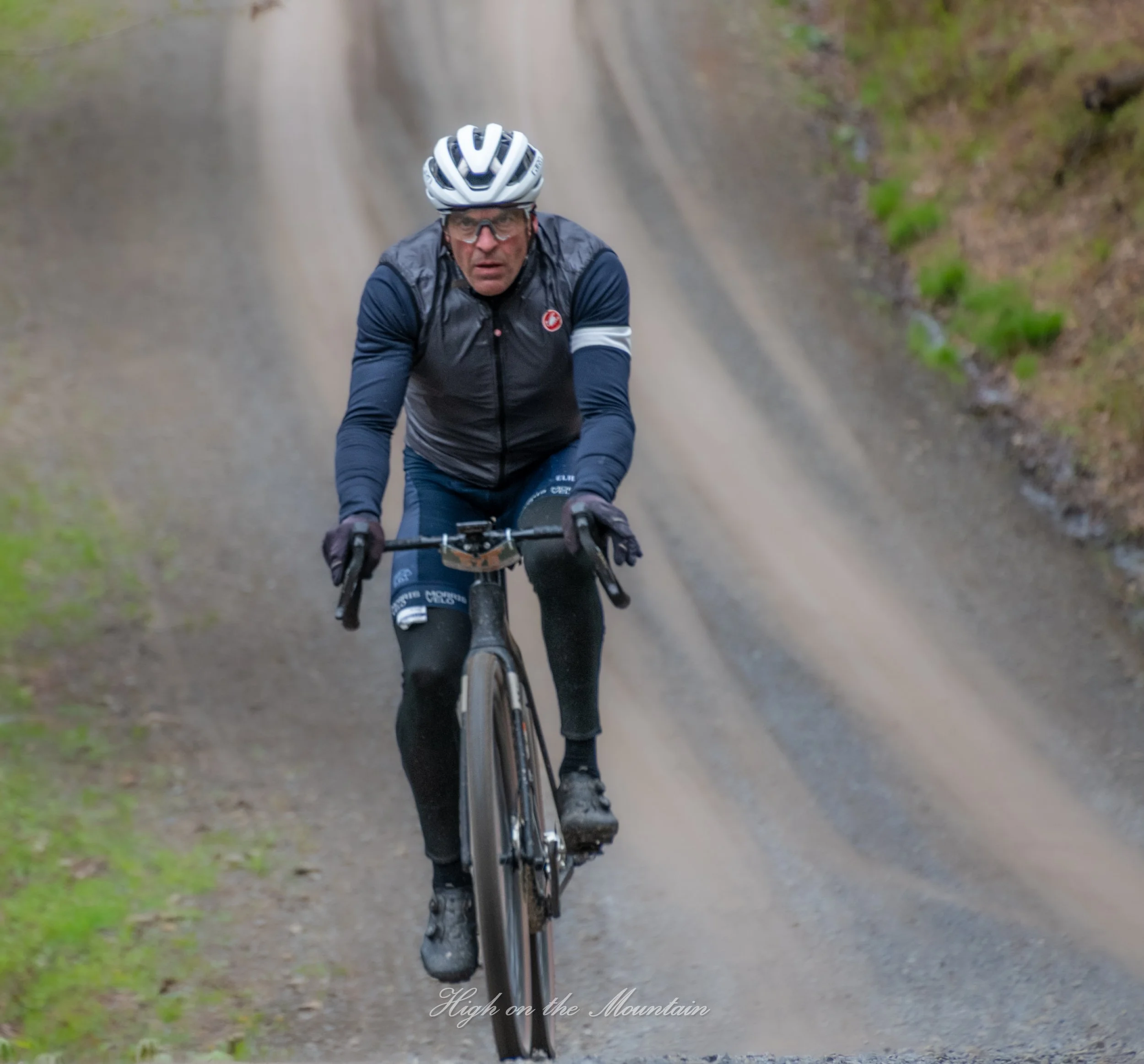 A man riding a mountain bike on a dirt trail in a natural outdoor setting. He is wearing a helmet, glasses, and a dark jacket. The scene appears to be in a forest or wooded area.