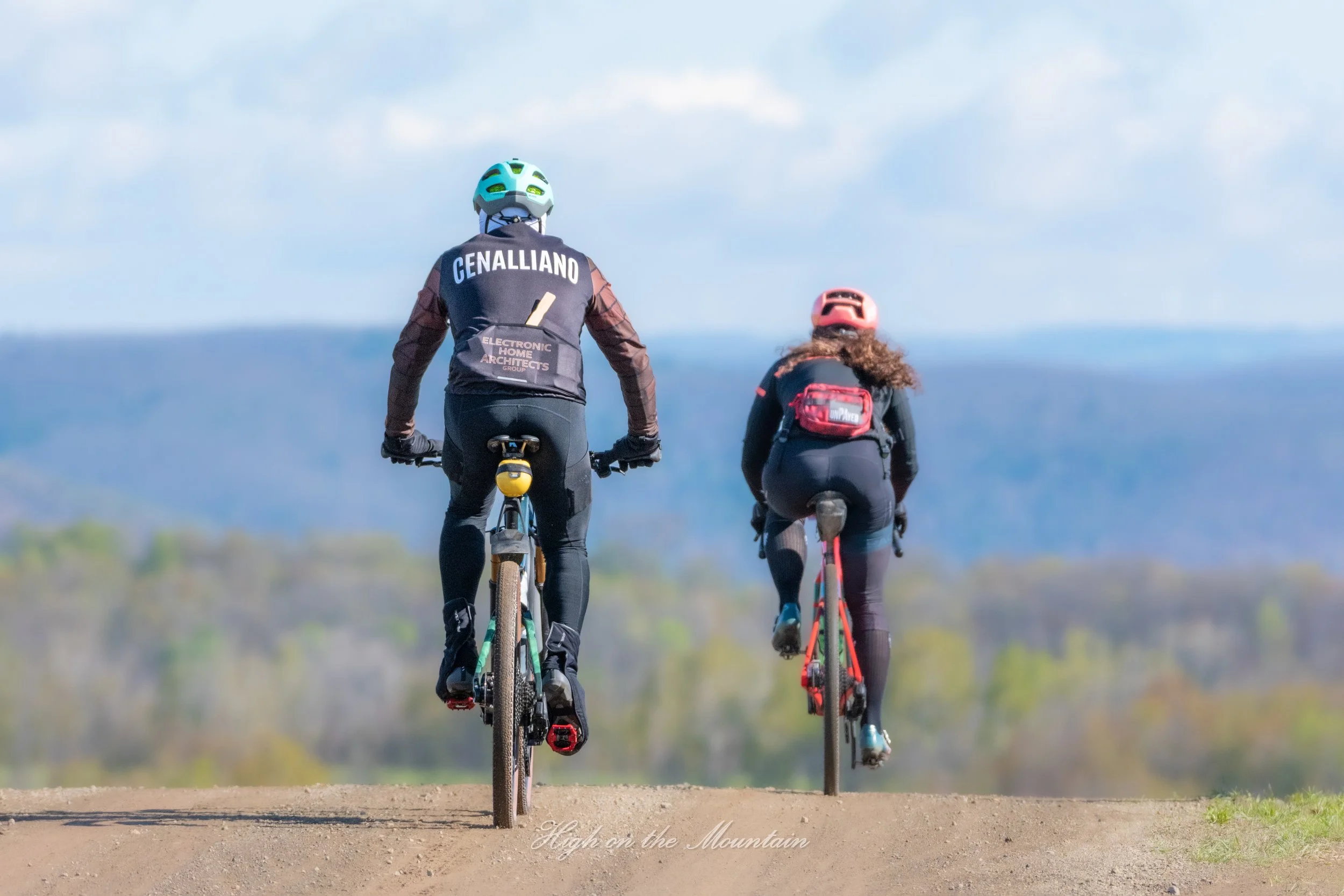 Two people riding bicycles on a dirt trail with a scenic mountain view under a partly cloudy sky.