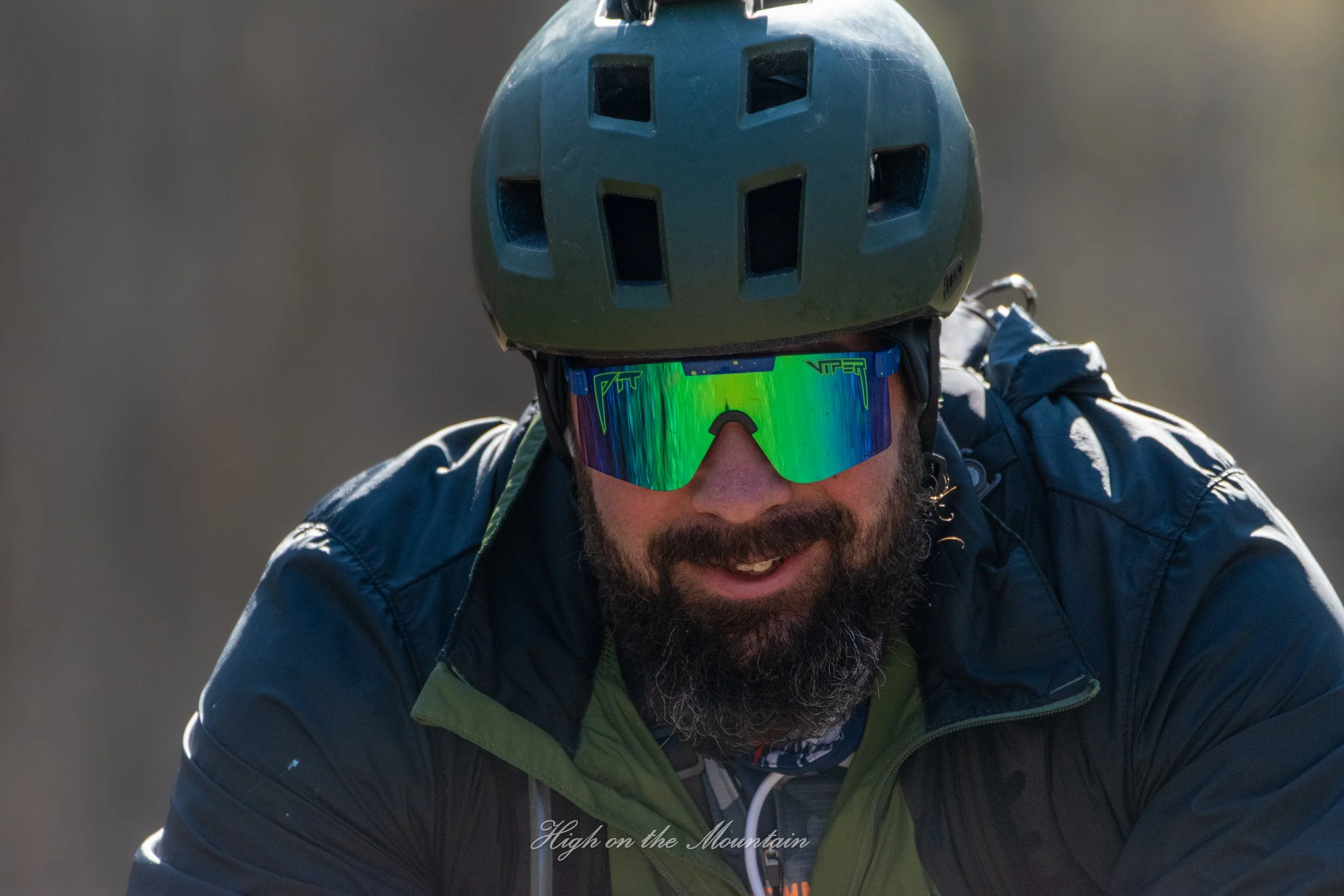A man wearing a black bicycle helmet, reflective sunglasses, a black jacket, and a green shirt is riding a bike on a mountain trail, smiling and looking ahead.