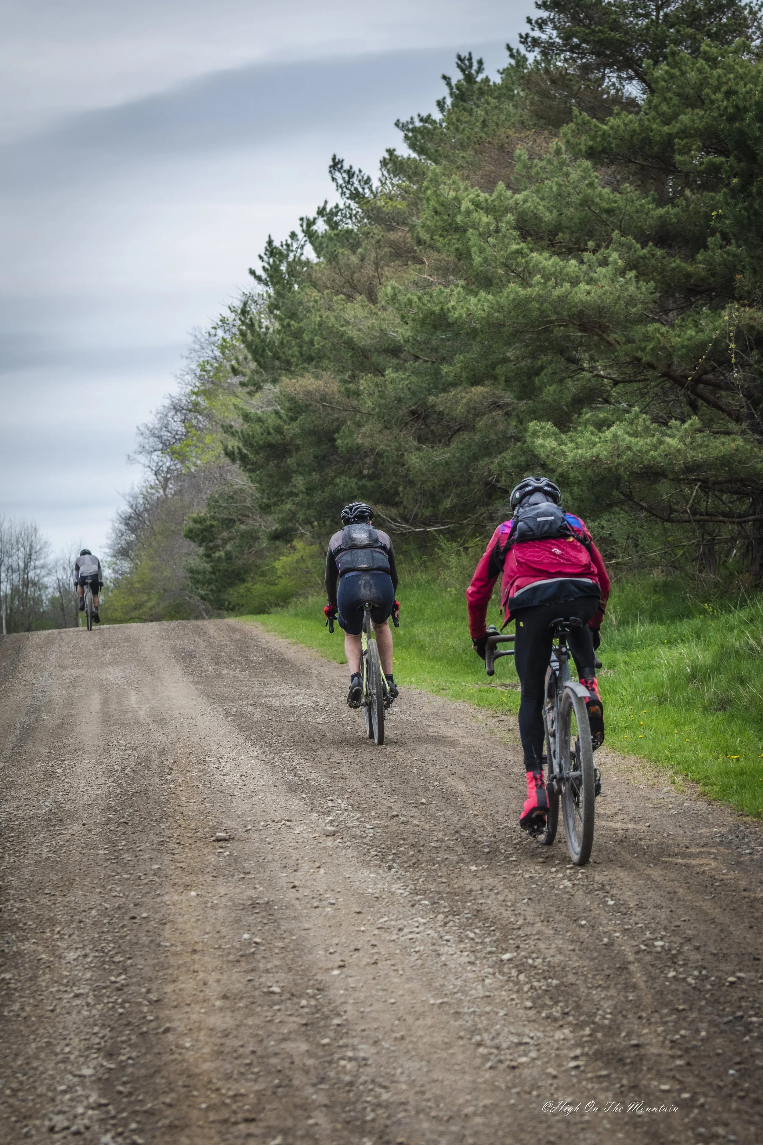 Three cyclists riding on a dirt path through a green wooded area under a cloudy sky.