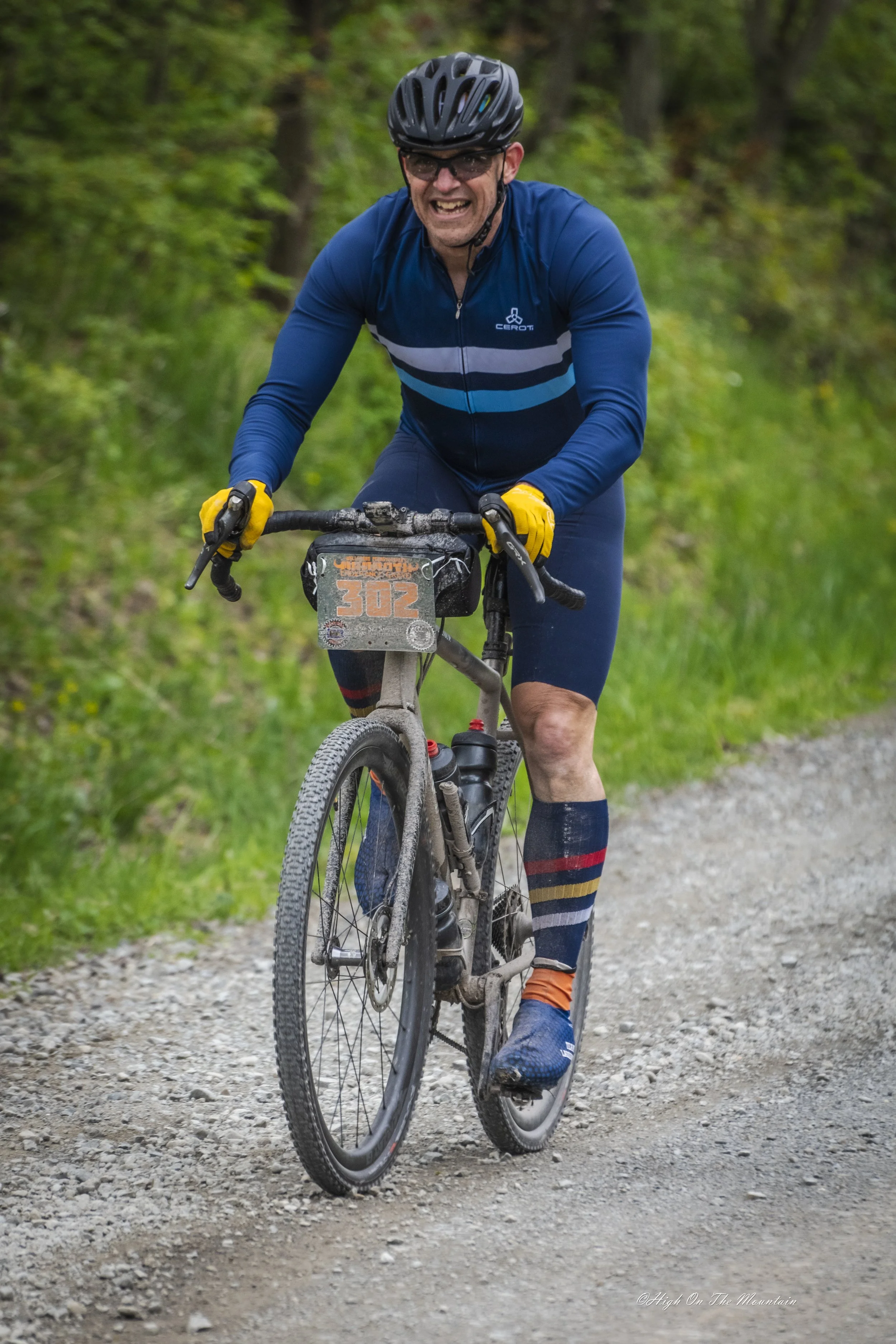 A male mountain biker riding on a gravel trail in a wooded area, wearing a black helmet, glasses, a blue cycling jacket, and yellow gloves, with trees and green foliage in the background.