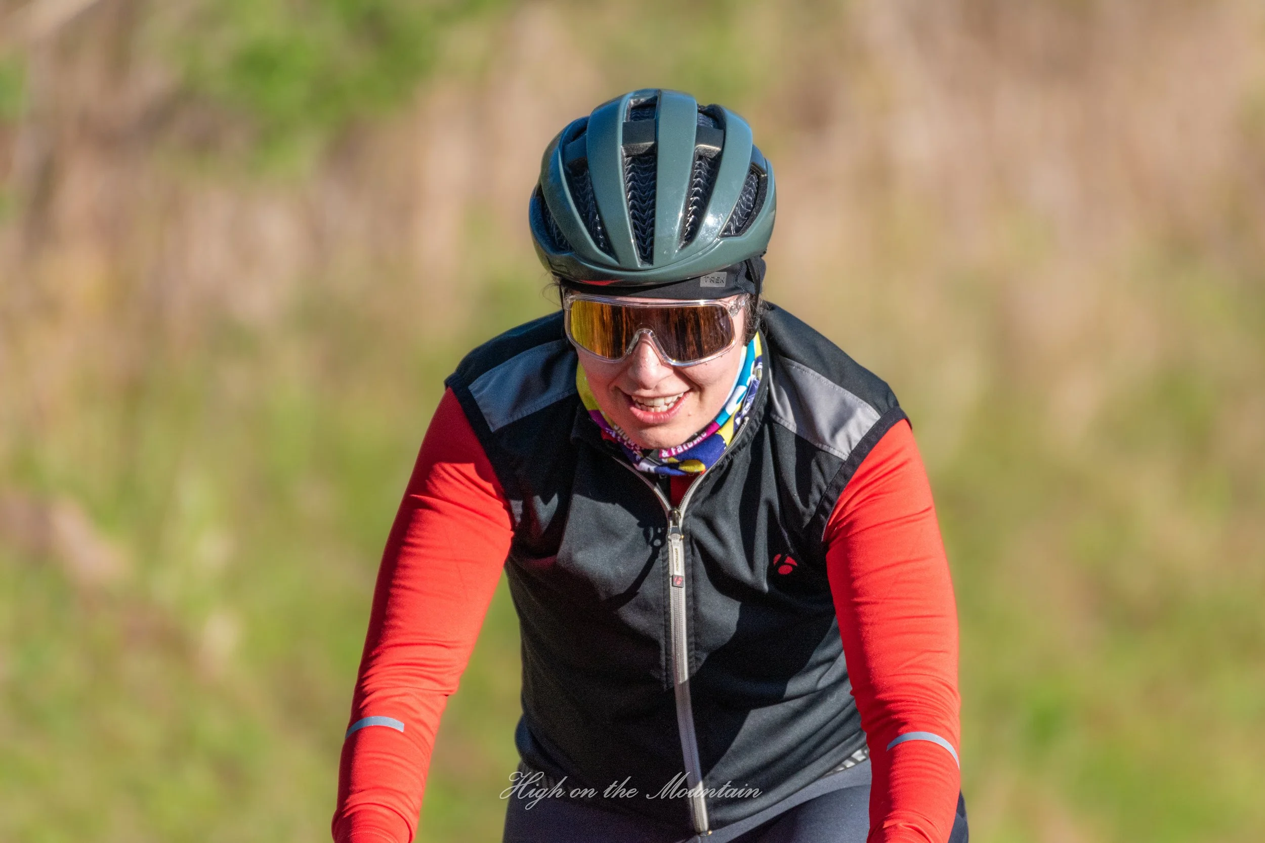 A person wearing a black helmet, sunglasses, and a black and red jacket riding a bike outdoors on a sunny day.
