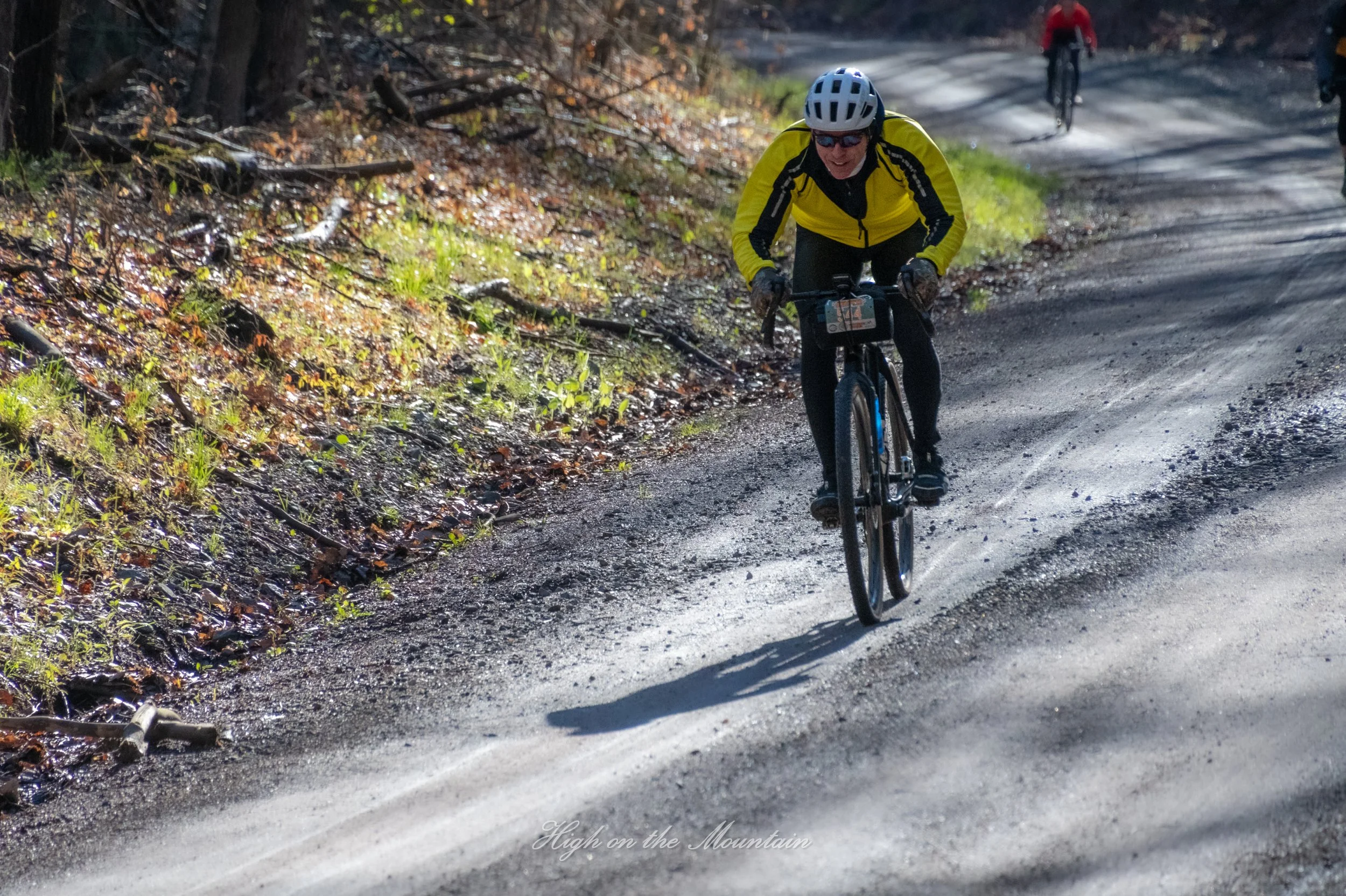 A person wearing a yellow jacket, black pants, helmet, and sunglasses riding a mountain bike on a dirt trail through a wooded area. Two other bikers are riding in the background.