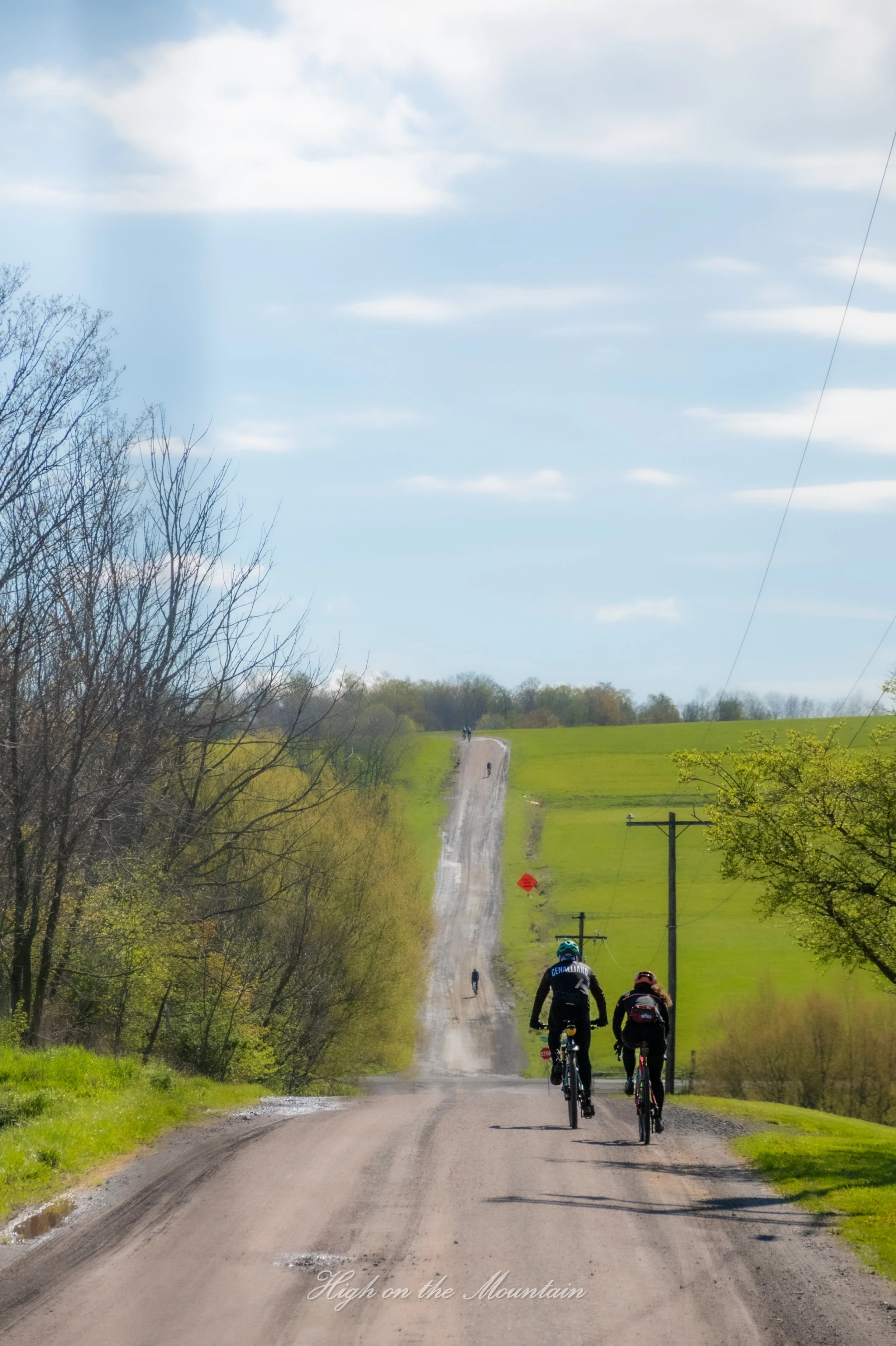 Cyclists riding down a rural dirt road through green fields with trees and hills in the background, under a partly cloudy sky, with the text 'High on the Mountain' at the bottom of the image.