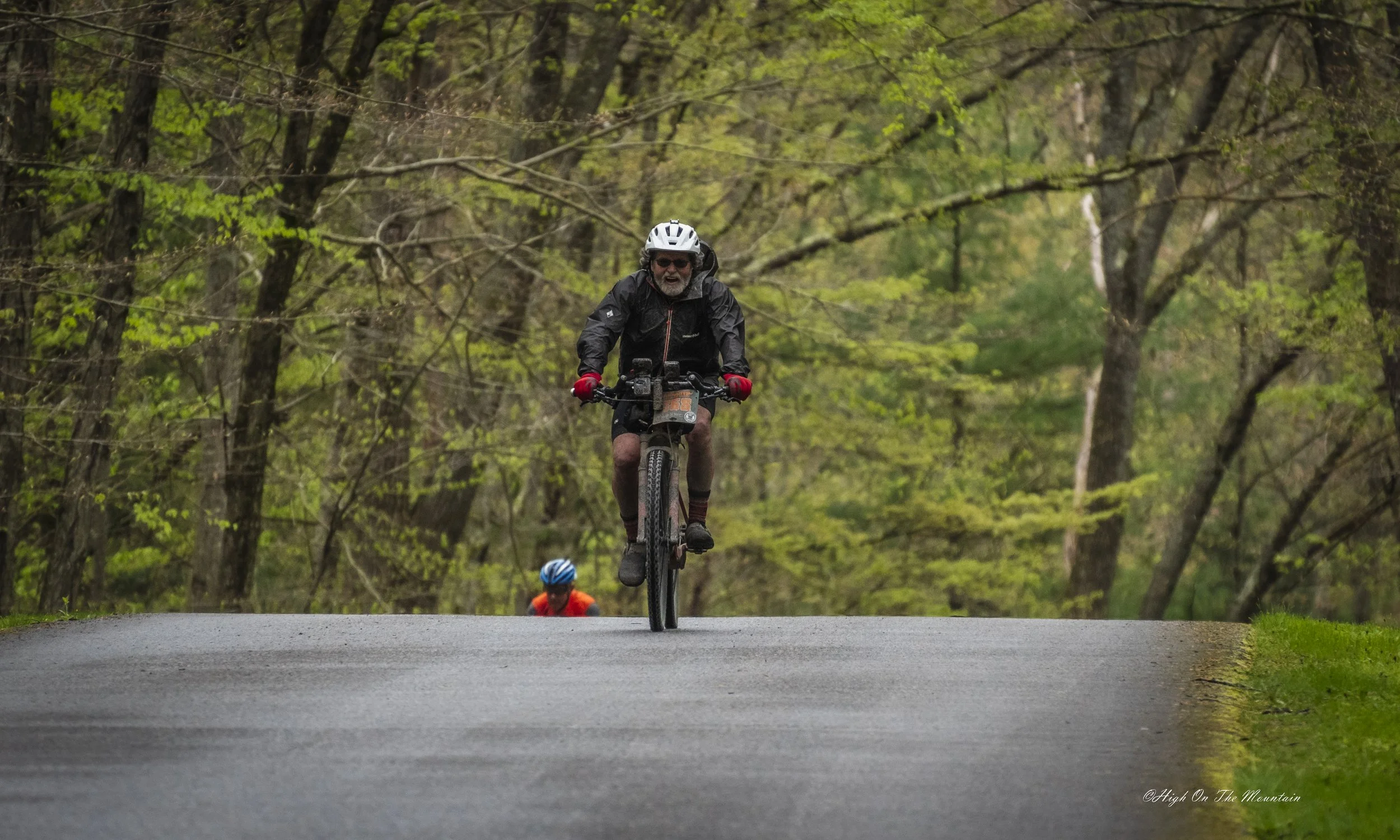 A man riding a bicycle on a forest trail with trees in the background, seen from the front, with a second cyclist partially visible behind him.