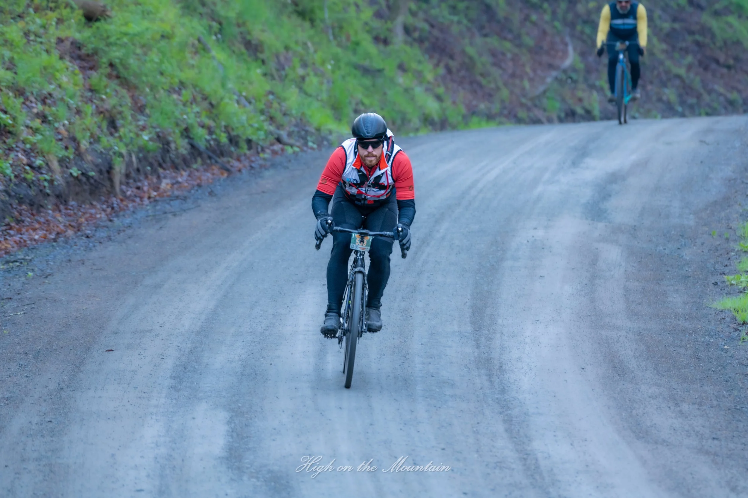 A man wearing a black helmet, sunglasses, and a red jacket rides a bicycle on a dirt mountain trail, with a forested hill on the side. Another cyclist is visible in the background.