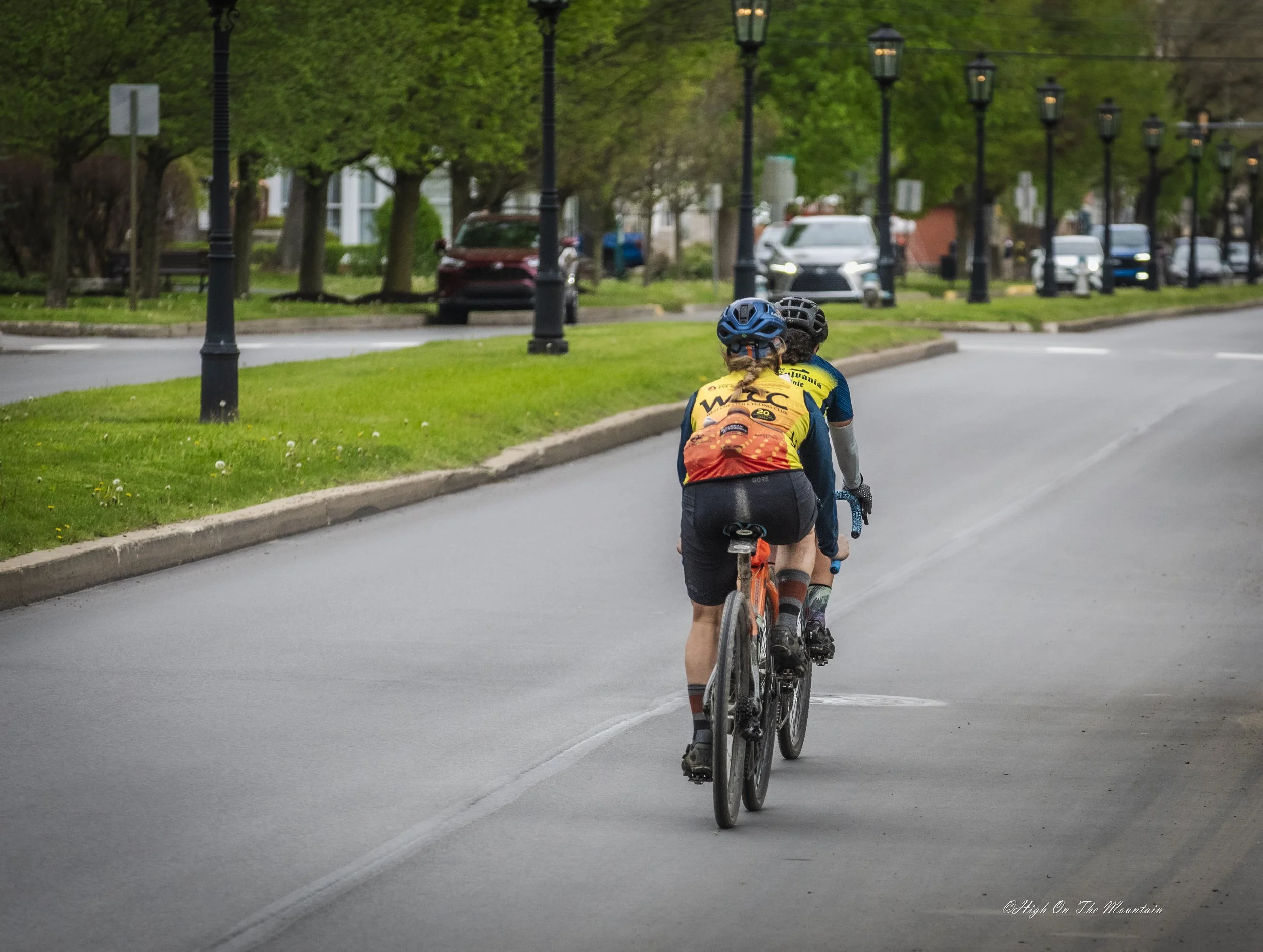 Two cyclists, wearing helmets and cycling jerseys, ride their bikes on a paved road in a park-like area with trees and street lamps lining the side of the road. There are parked cars and a building in the background.