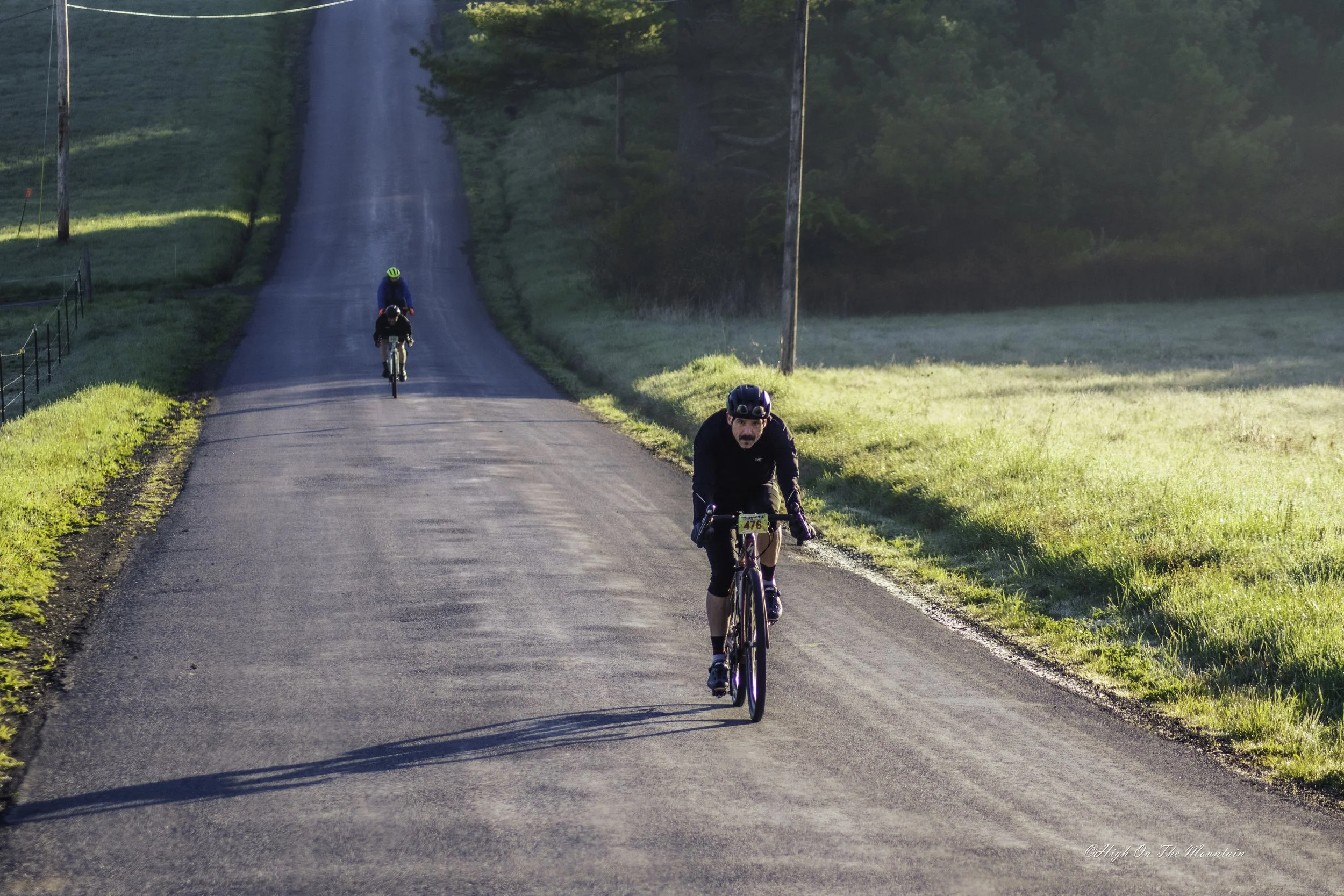 Three cyclists riding on a rural road surrounded by green grass and trees, with one cyclist in the foreground and two further back, during what appears to be early morning or late afternoon.