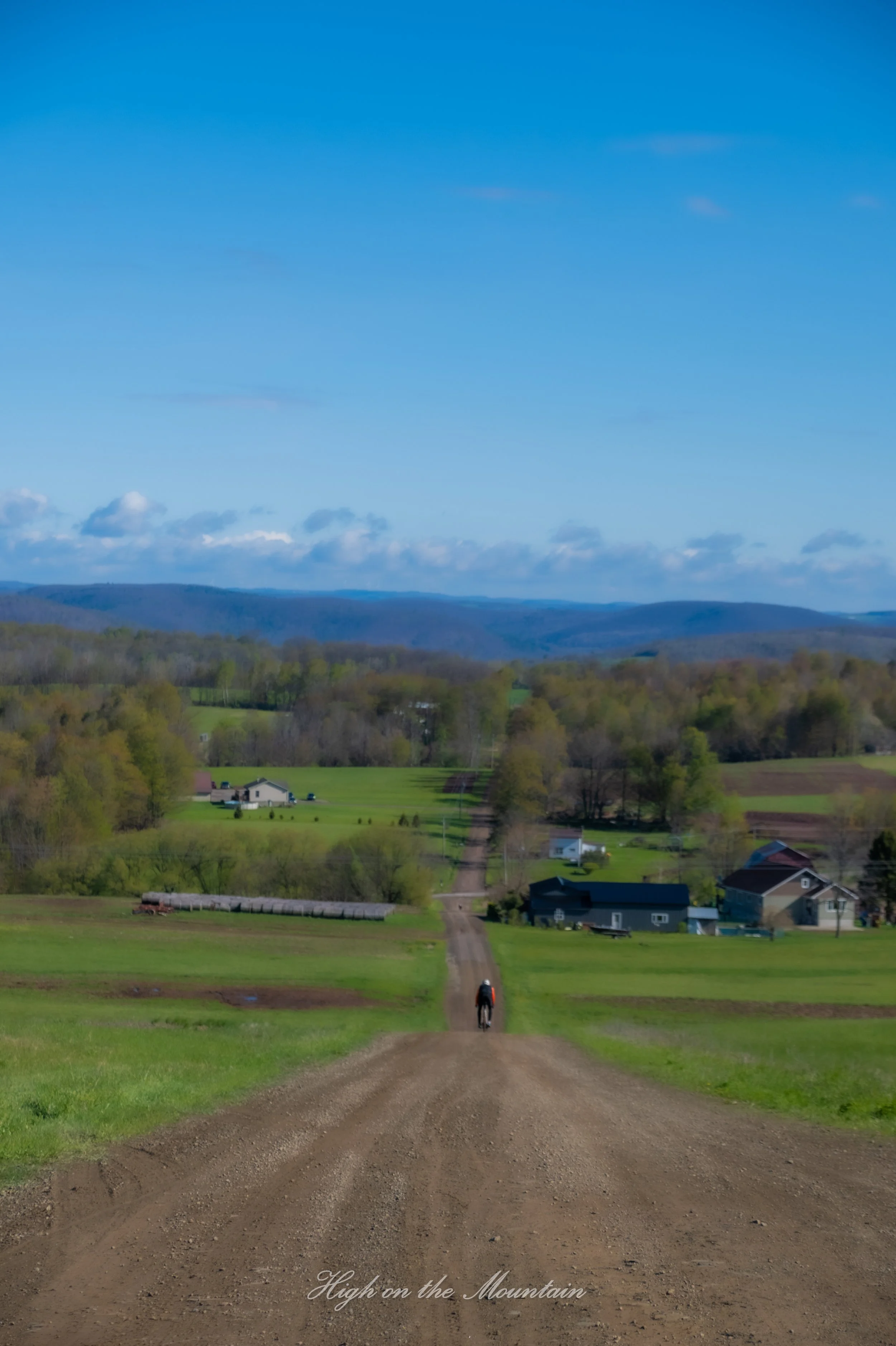 A rural landscape with a dirt road leading toward houses and green fields, rolling hills in the distance, and a blue sky with few clouds. A person is riding a bicycle on the road, and the scene is captioned 'High on the Mountain'.