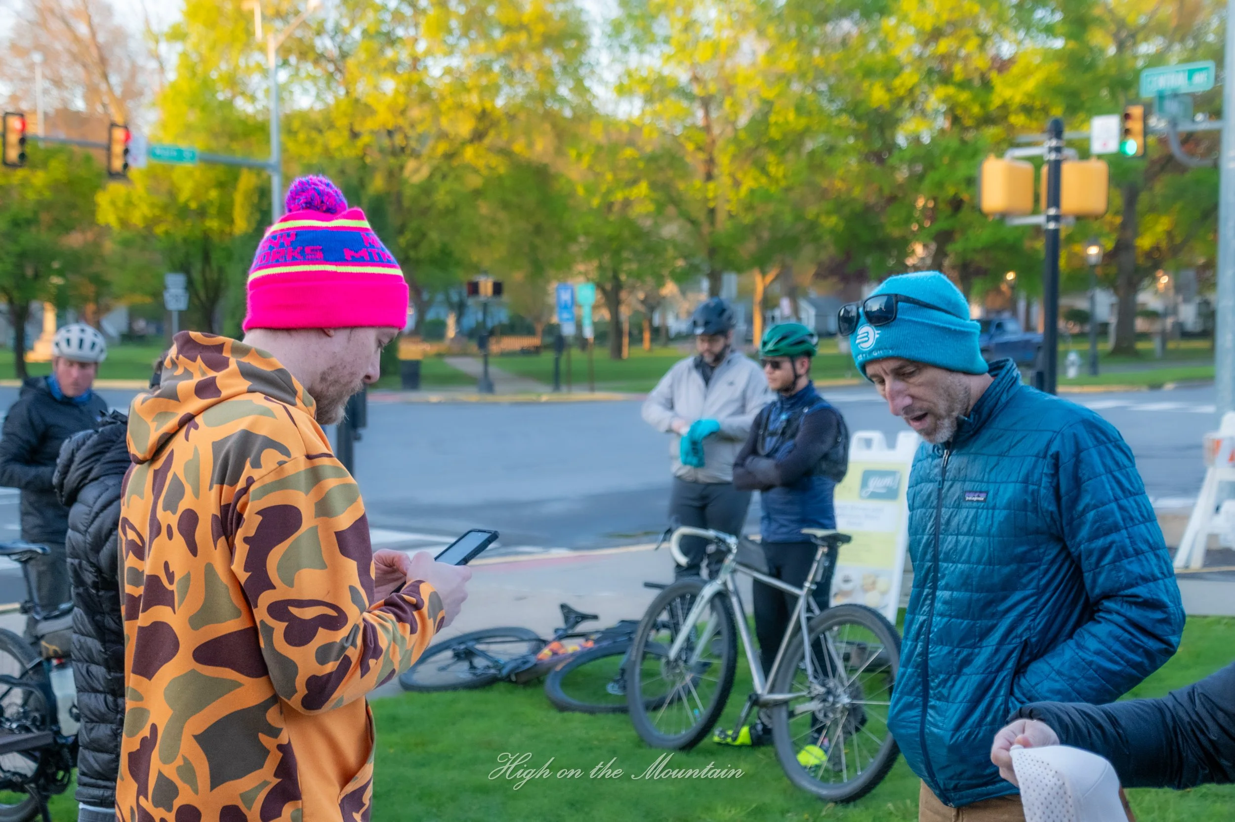 Group of people gathered outdoors near bicycles, some wearing helmets and jackets, with trees and traffic lights in the background.