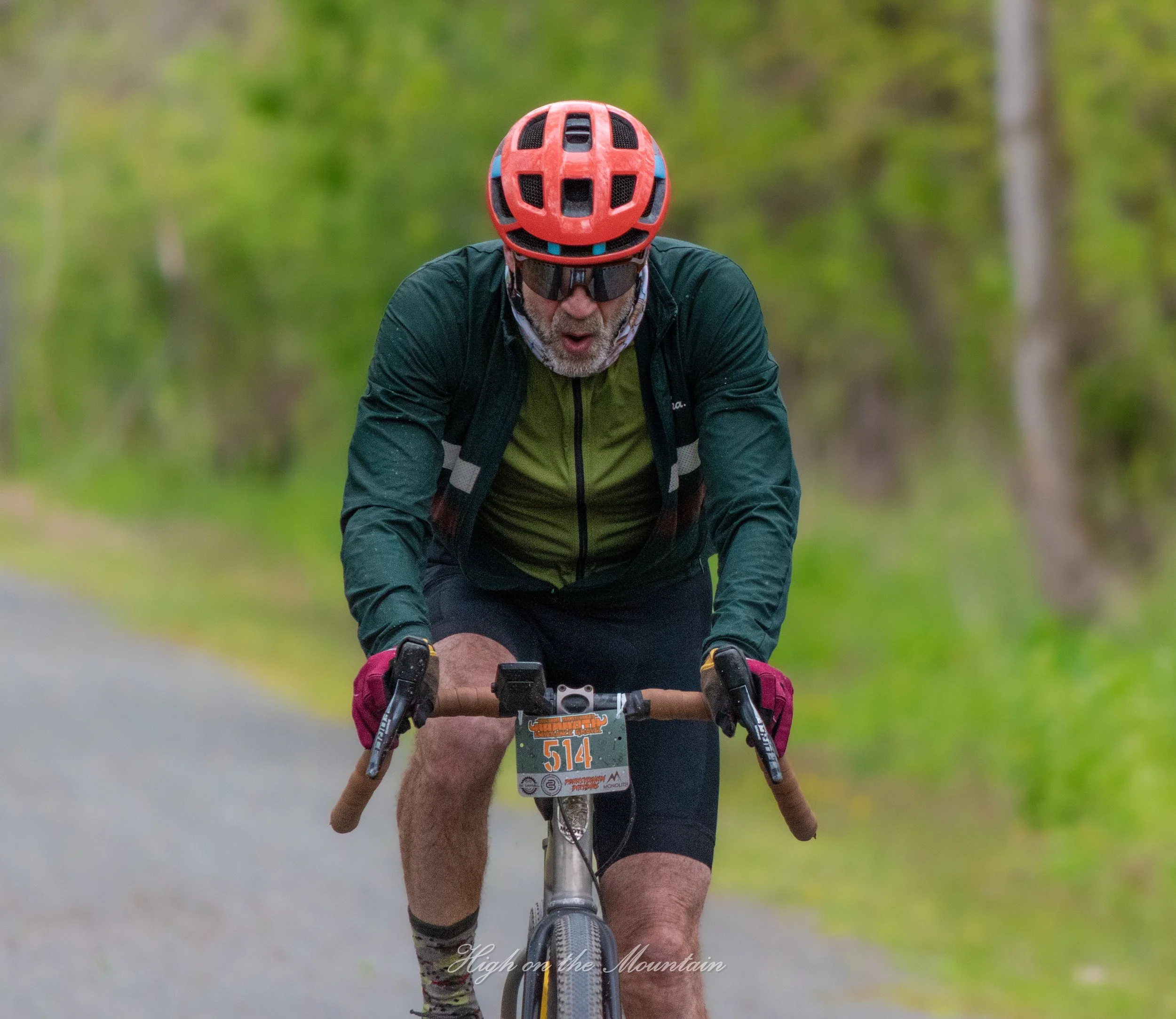 A man wearing a red helmet and sunglasses riding a mountain bike on a trail surrounded by green trees.