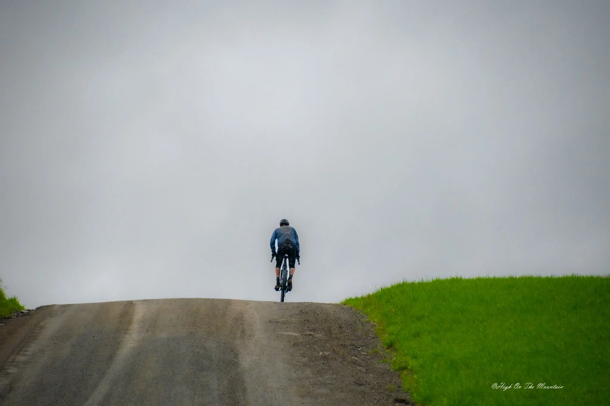 A person riding a bicycle up a dirt road surrounded by green grass on both sides under an overcast sky.
