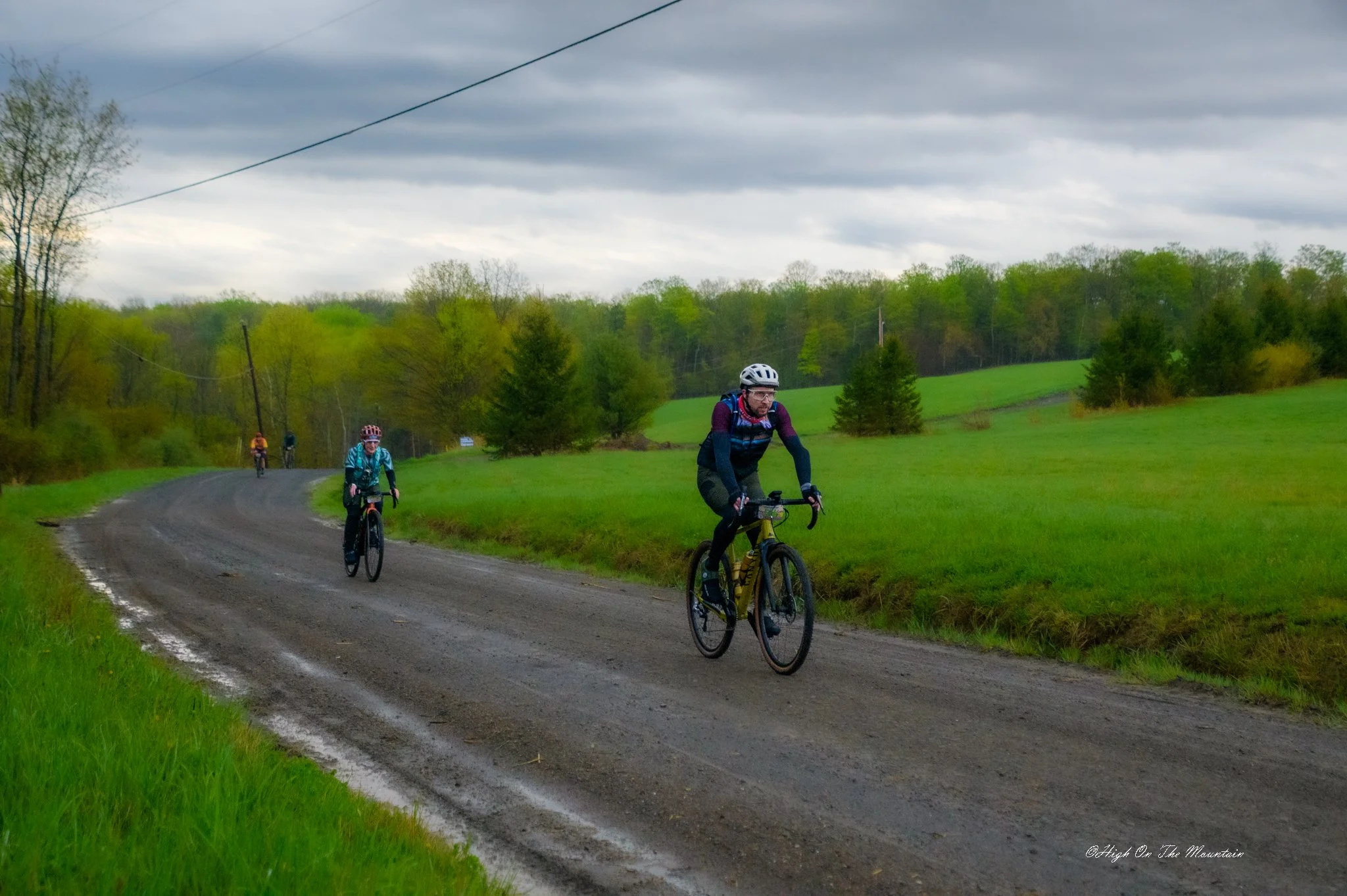 Three cyclists riding on a rural dirt road surrounded by green trees and grass under a cloudy sky.