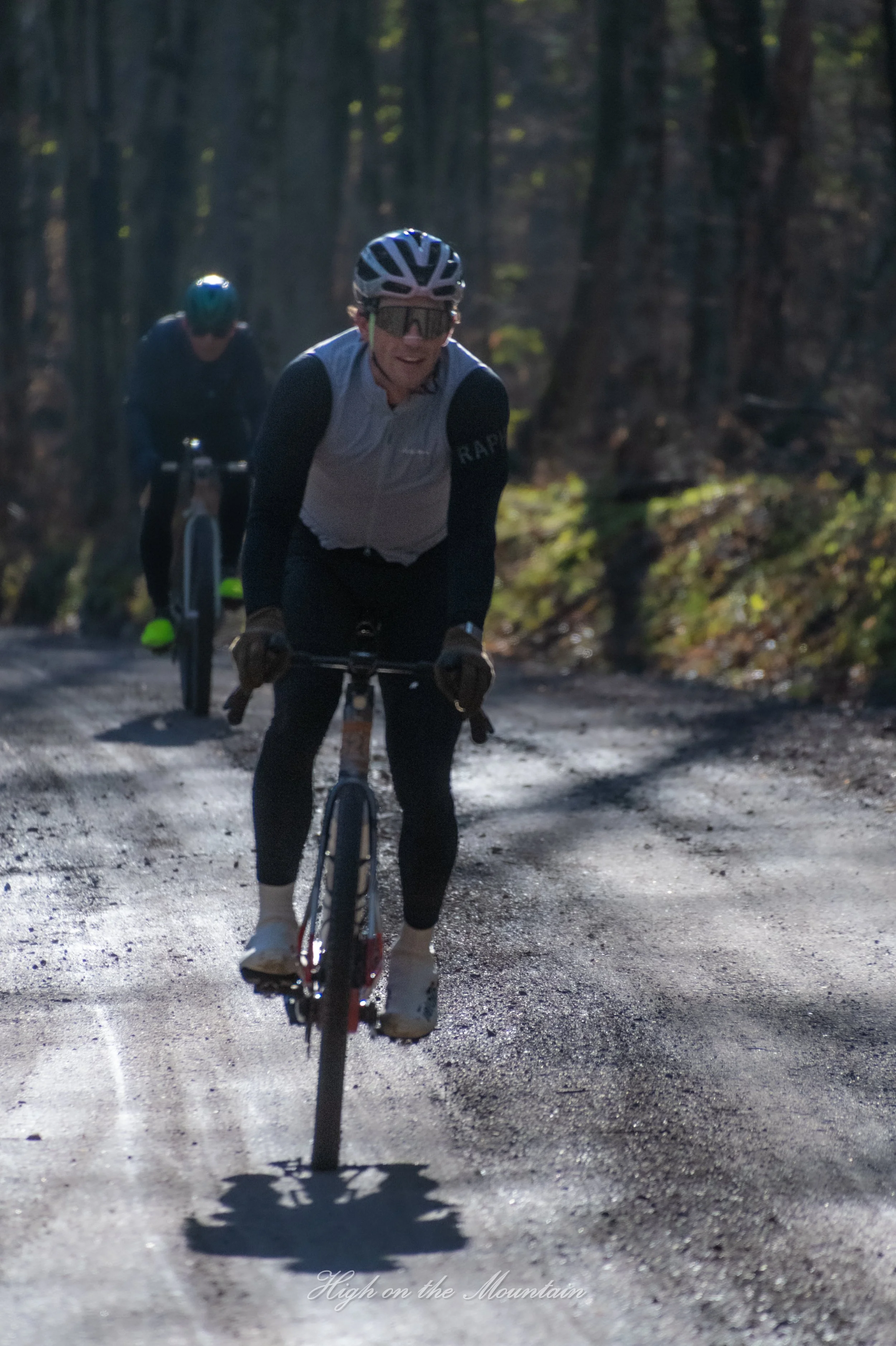 Two cyclists riding mountain bikes on a dirt trail through a wooded area, sun shining through trees.