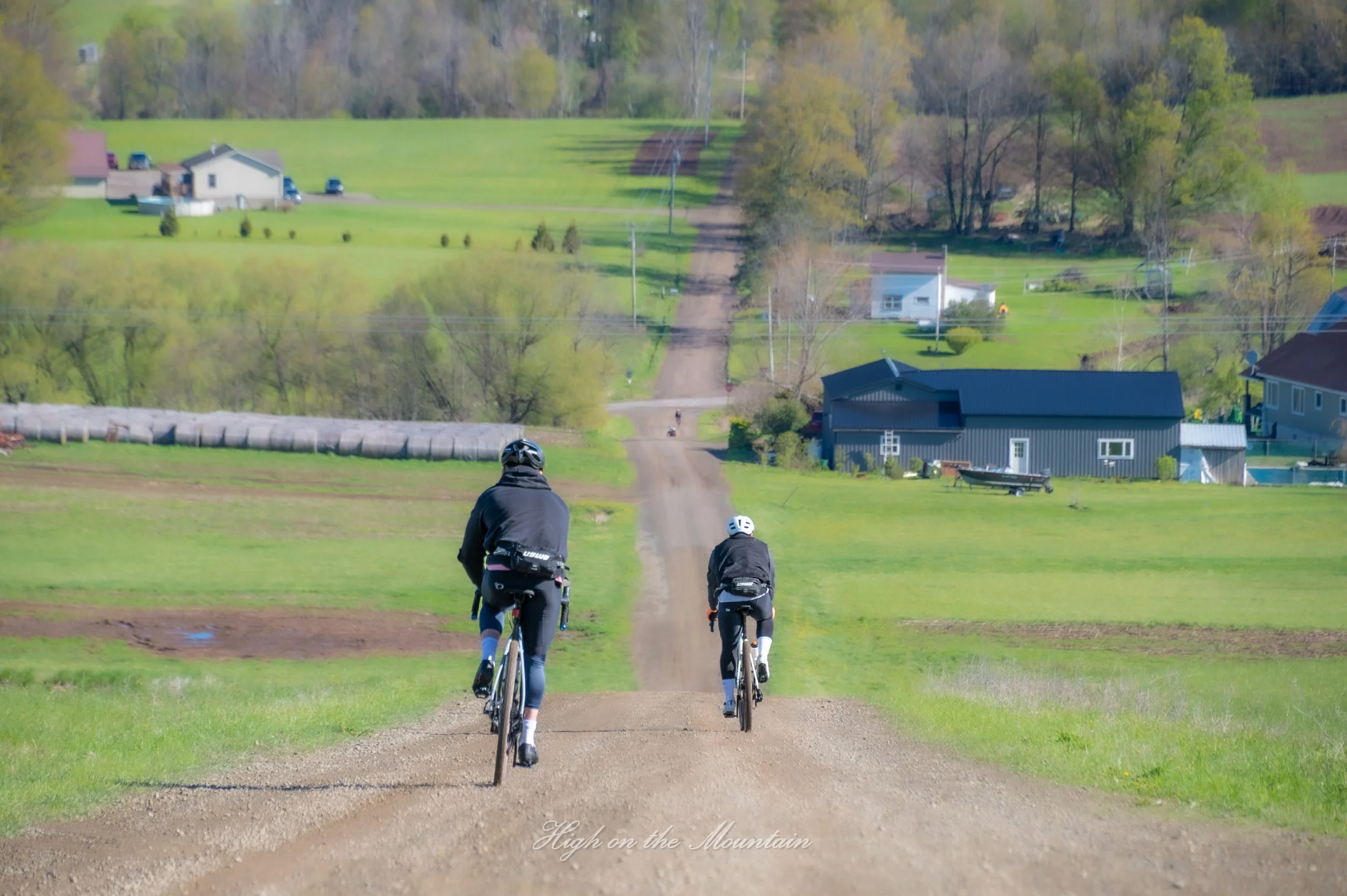 Three cyclists riding downhill on a dirt trail through a green rural landscape with houses and trees in the background.