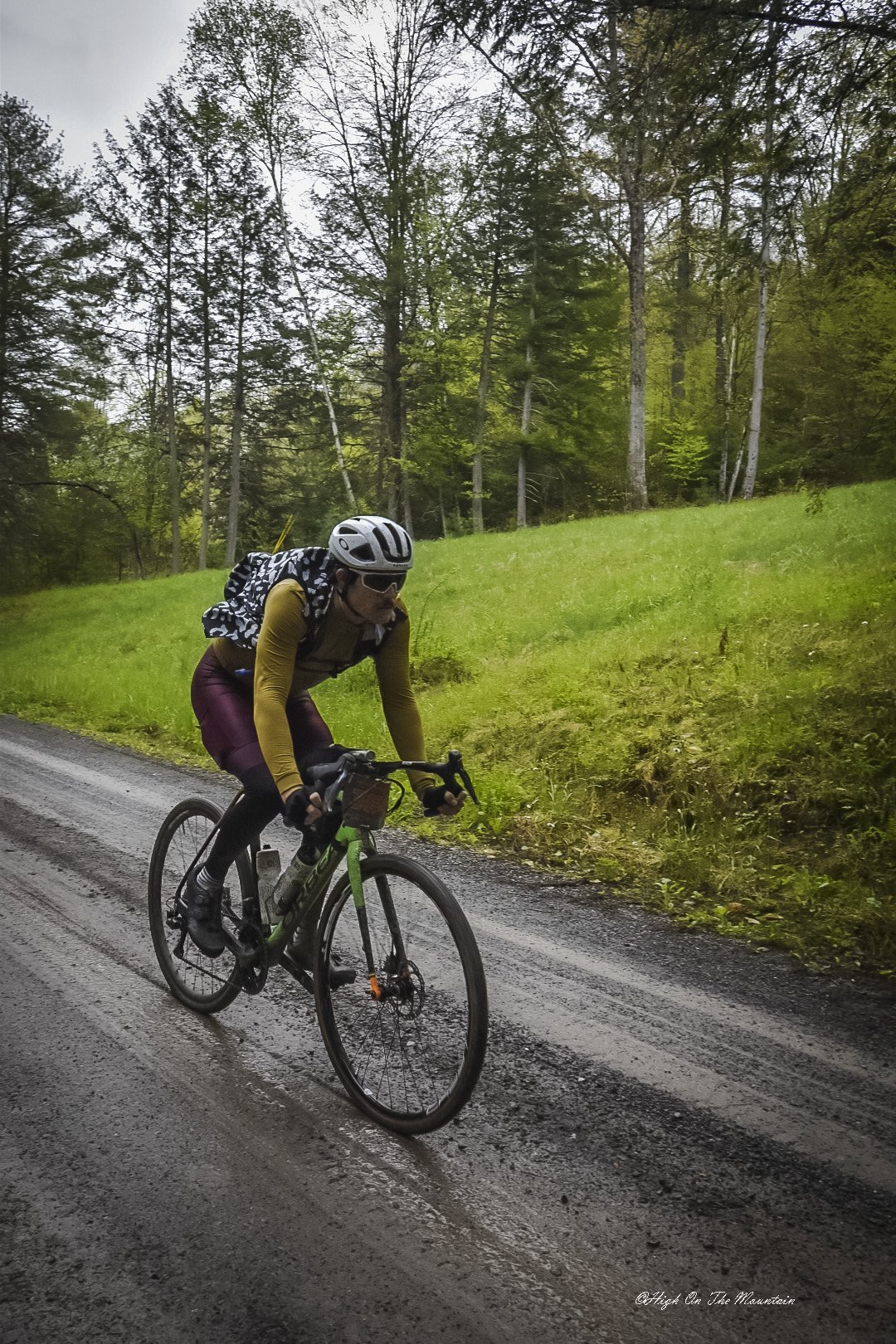 Cyclist wearing a helmet, sunglasses, yellow long sleeves, and maroon pants riding a mountain bike on a wet gravel road through a forested area.