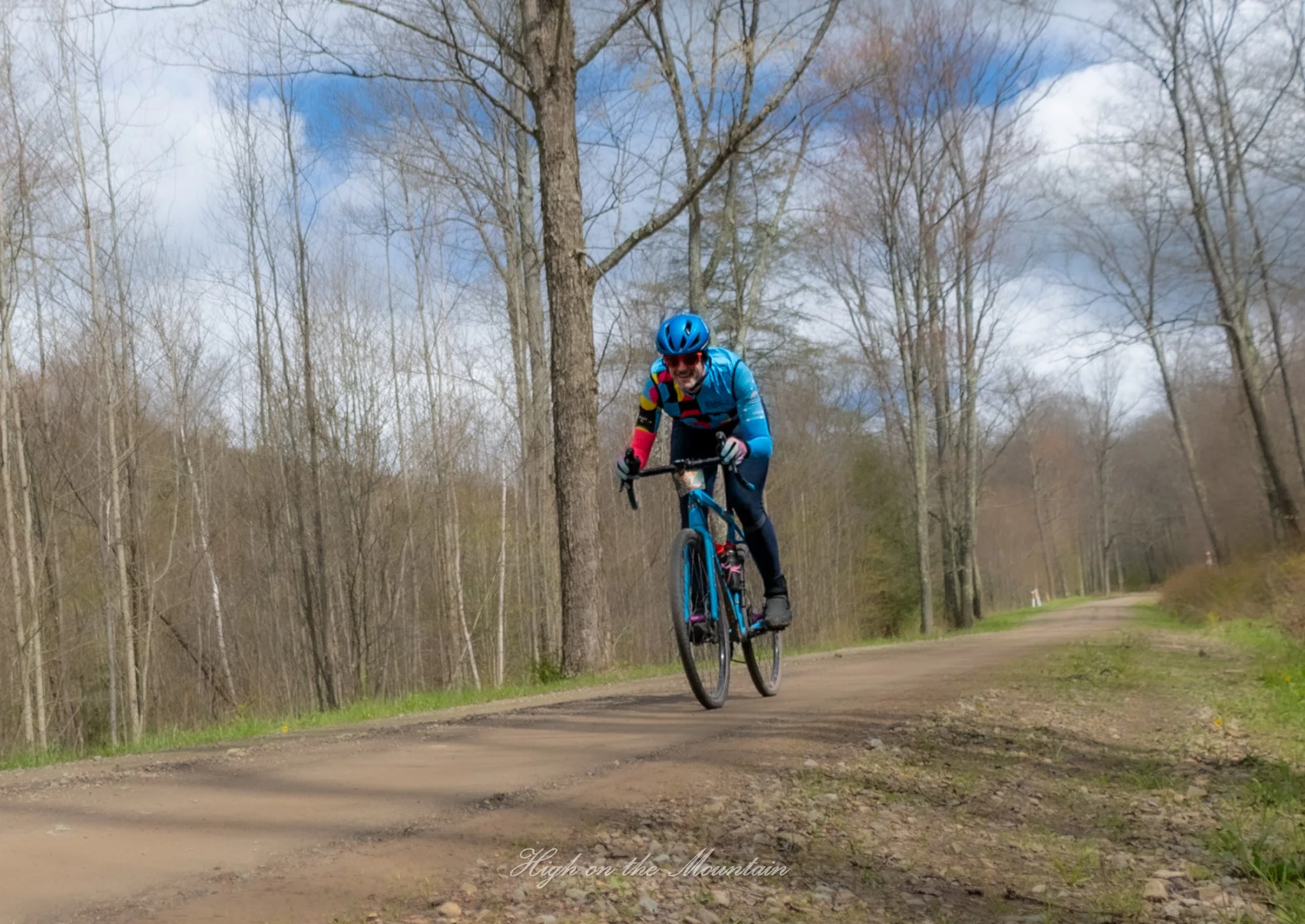A person wearing a blue helmet and colorful cycling gear riding a mountain bike on a dirt trail through a wooded area with leafless trees and a partly cloudy sky.