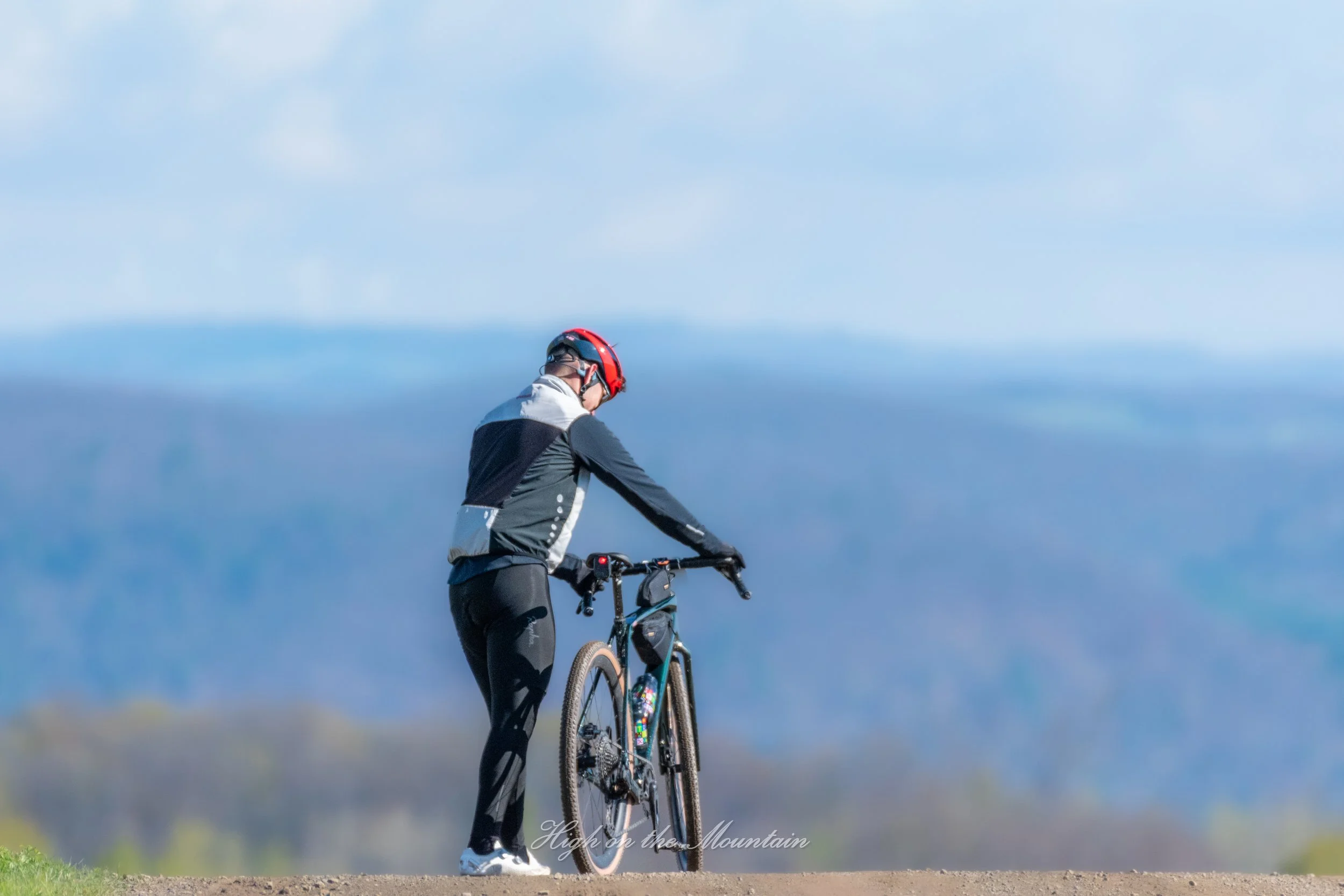 Person in cycling gear riding a mountain bike on a trail with mountainous landscape and sky in the background.