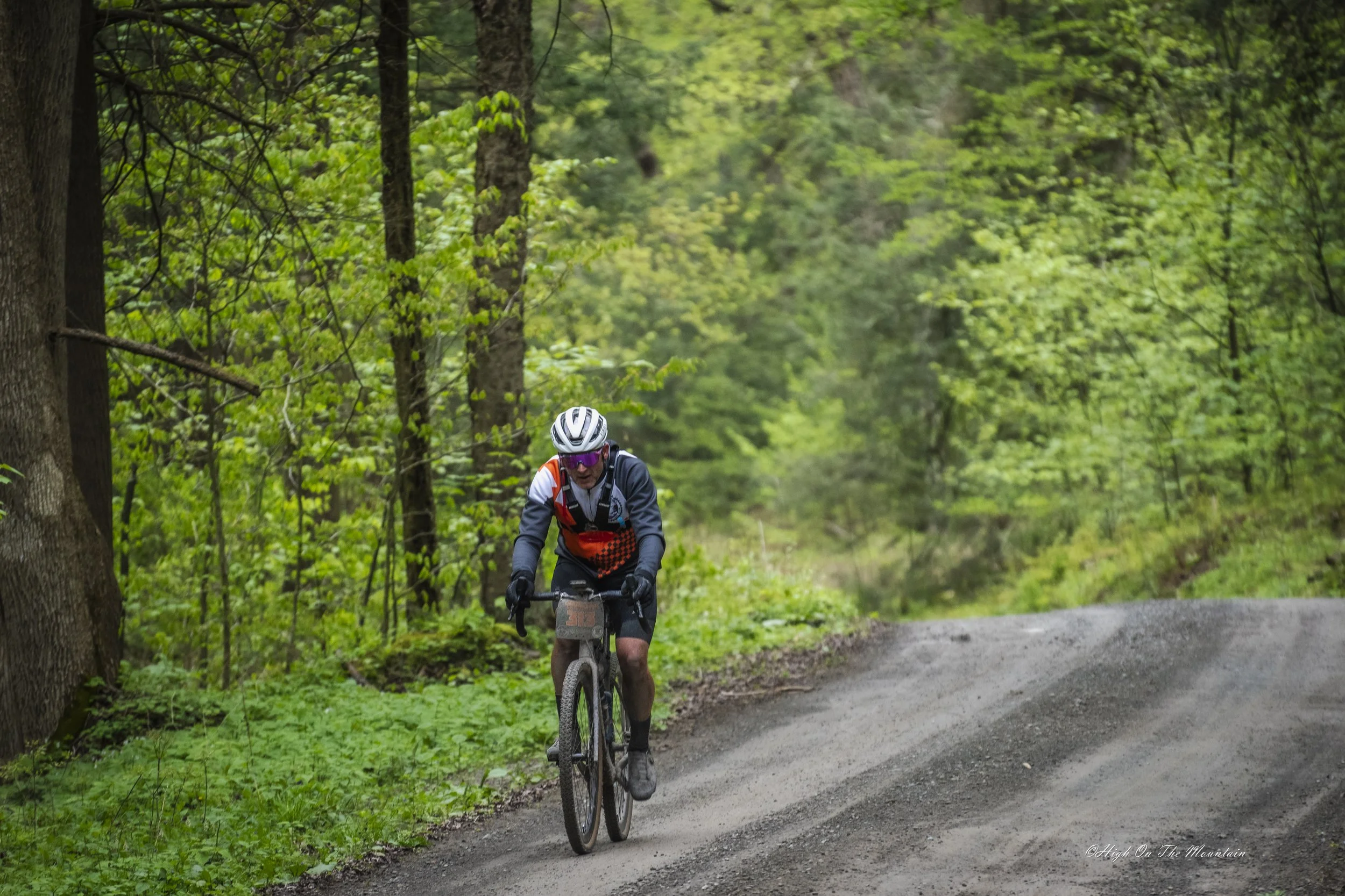 A mountain biker in a gray and orange outfit and helmet riding on a dirt trail through a lush green forest.