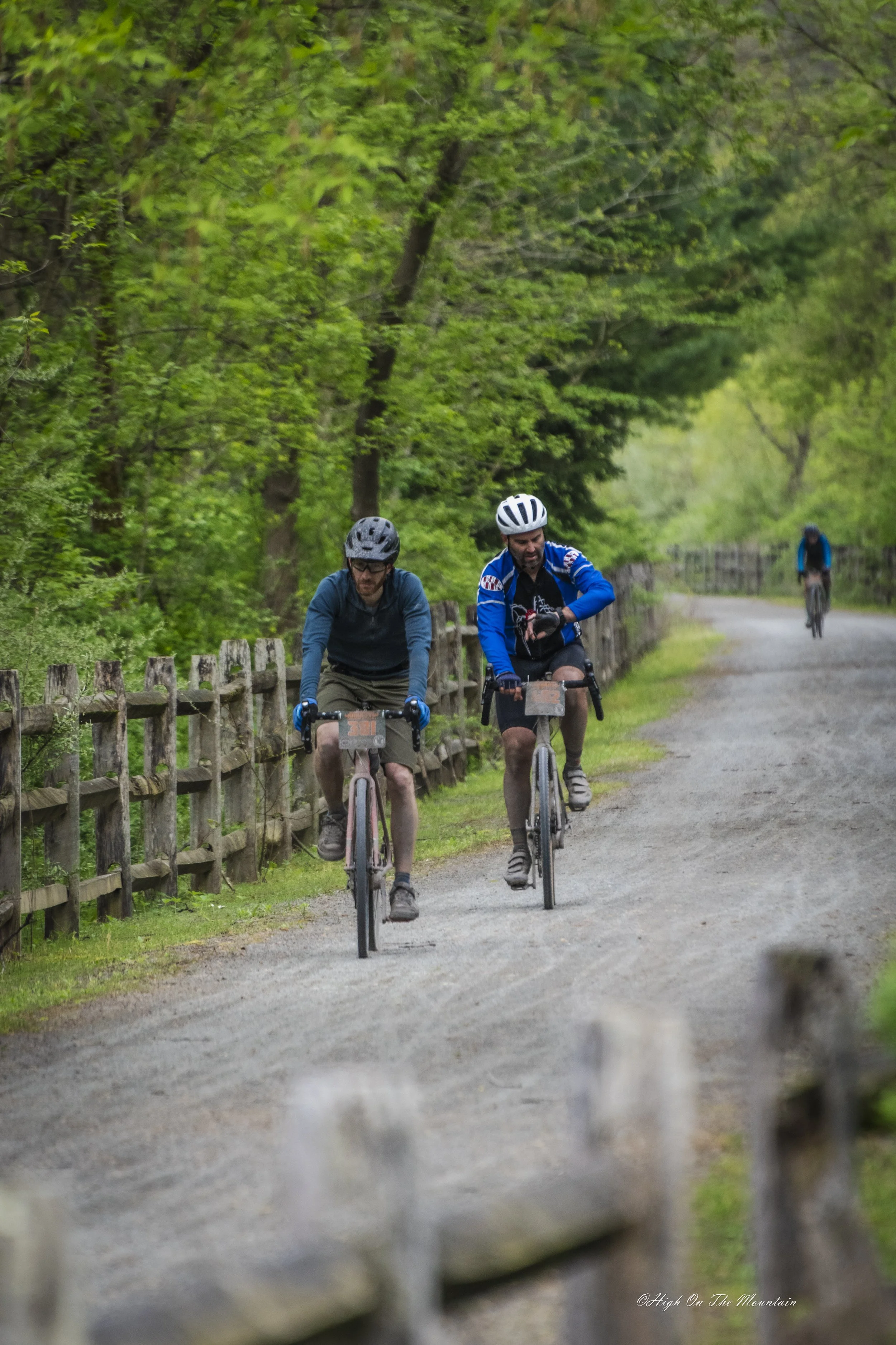 Two men riding mountain bikes on a dirt trail surrounded by green trees, with a wooden fence on the side of the trail in a forested area.