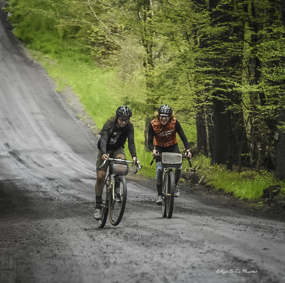 Two people riding mountain bikes on a dirt trail through a lush green forest, both wearing helmets and outdoor clothing.