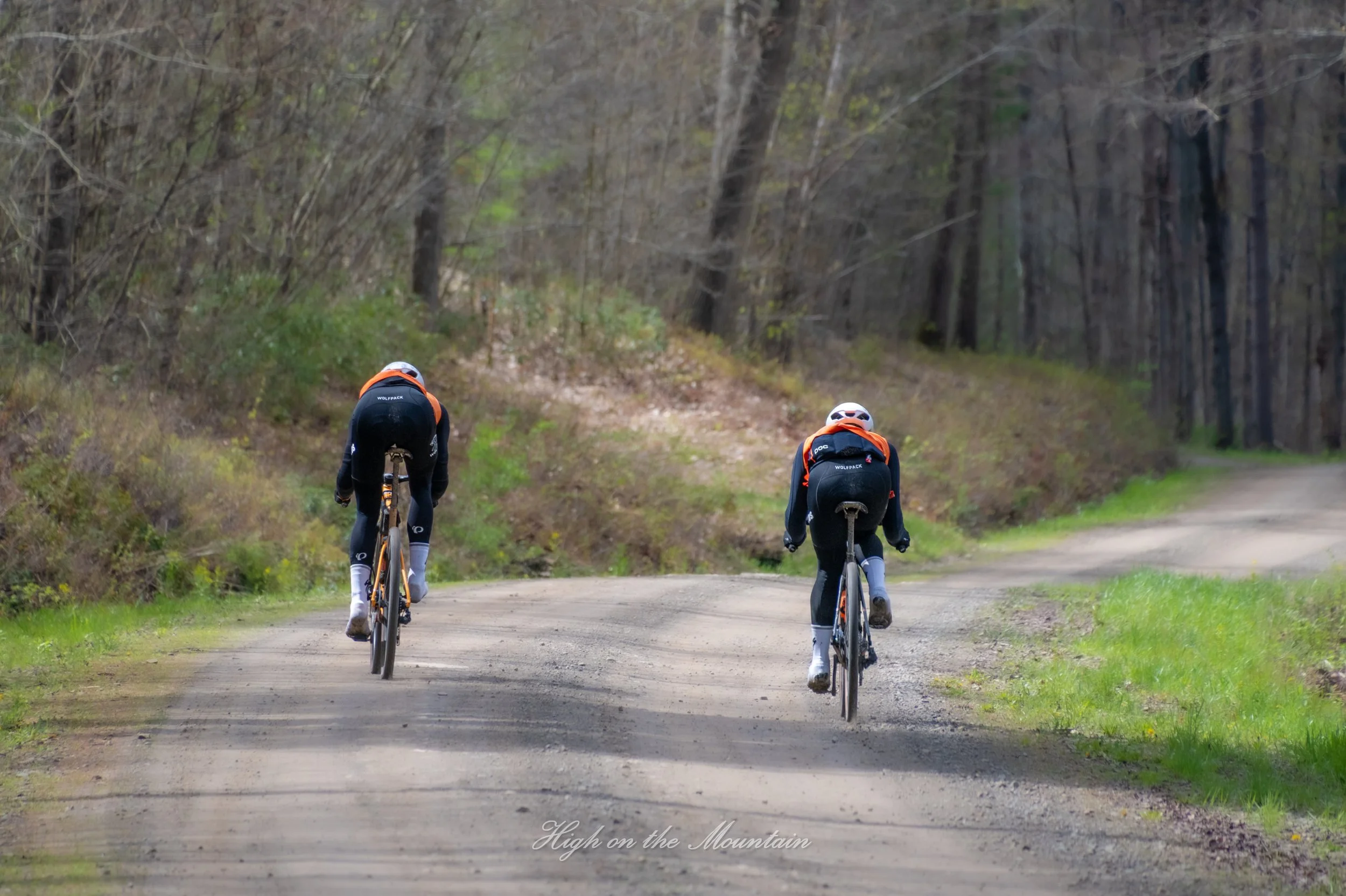 Two cyclists riding on a dirt trail through a forested area with leafless trees and sparse green foliage.