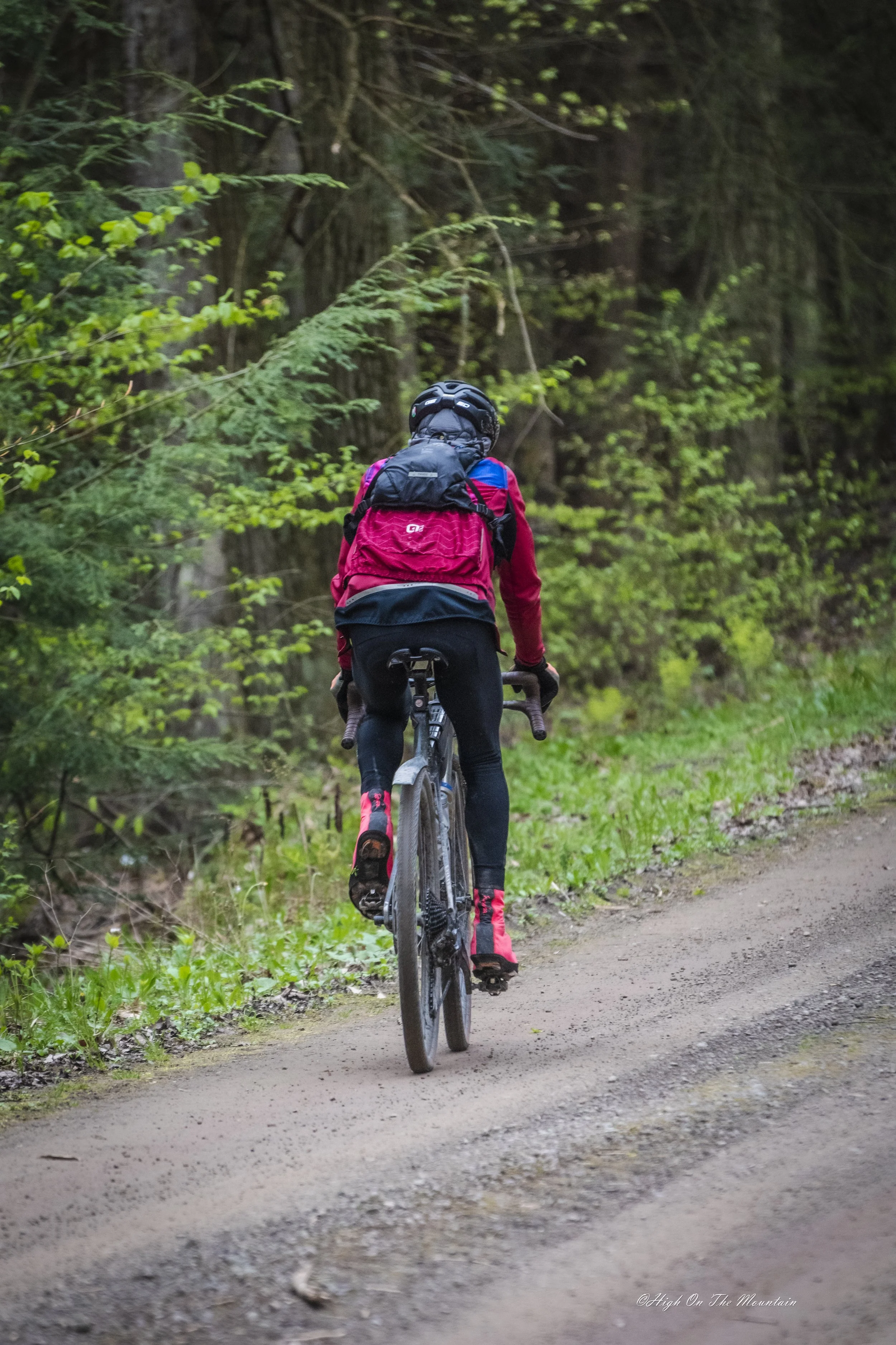Person riding a mountain bike on a dirt trail through a lush green forest, wearing a helmet, red jacket, and backpack.