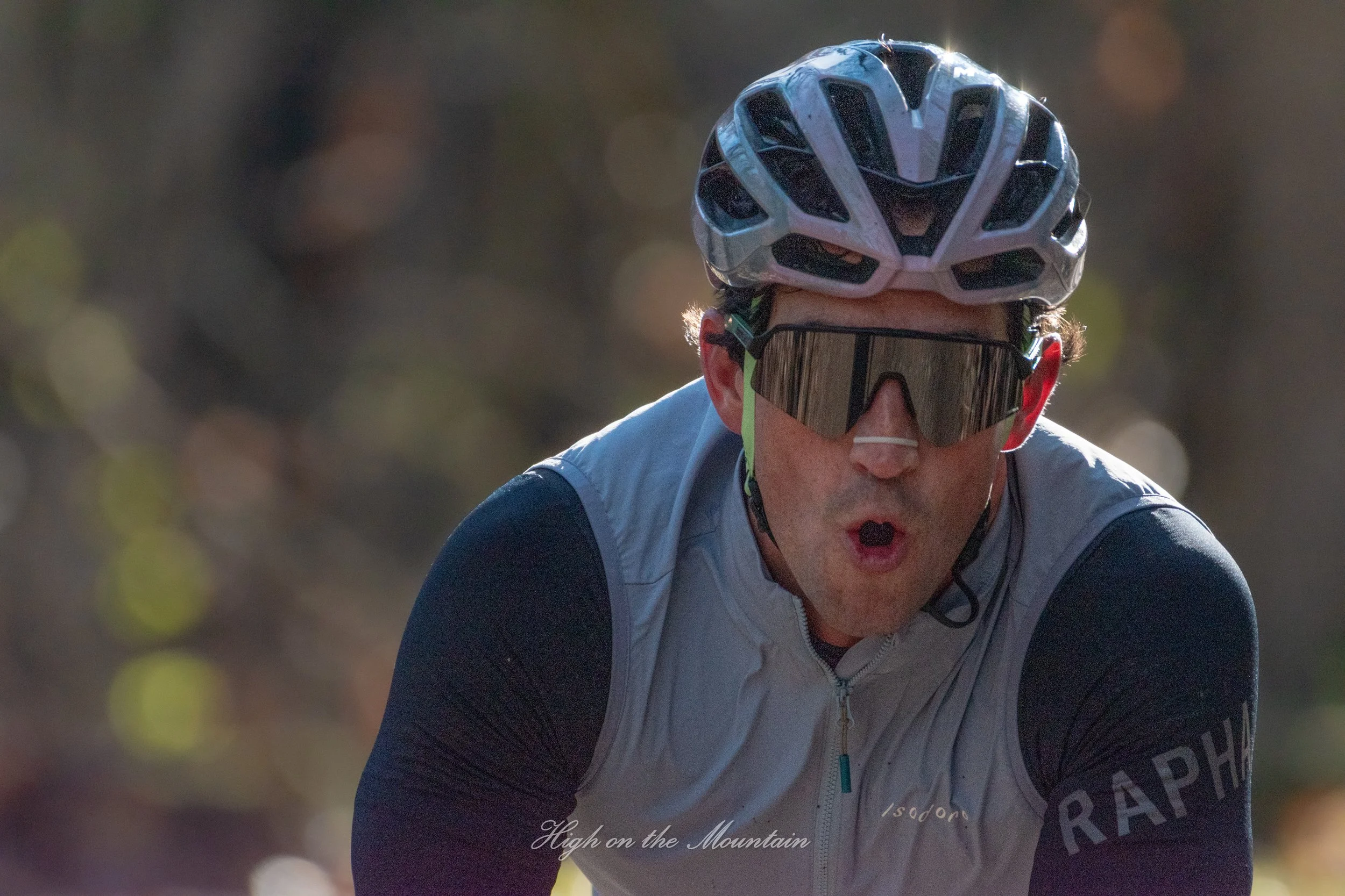 Male cyclist wearing a helmet and sunglasses cycling outdoors amidst blurred foliage with sunlight filtering through, wearing a primarily white and black cycling jersey.