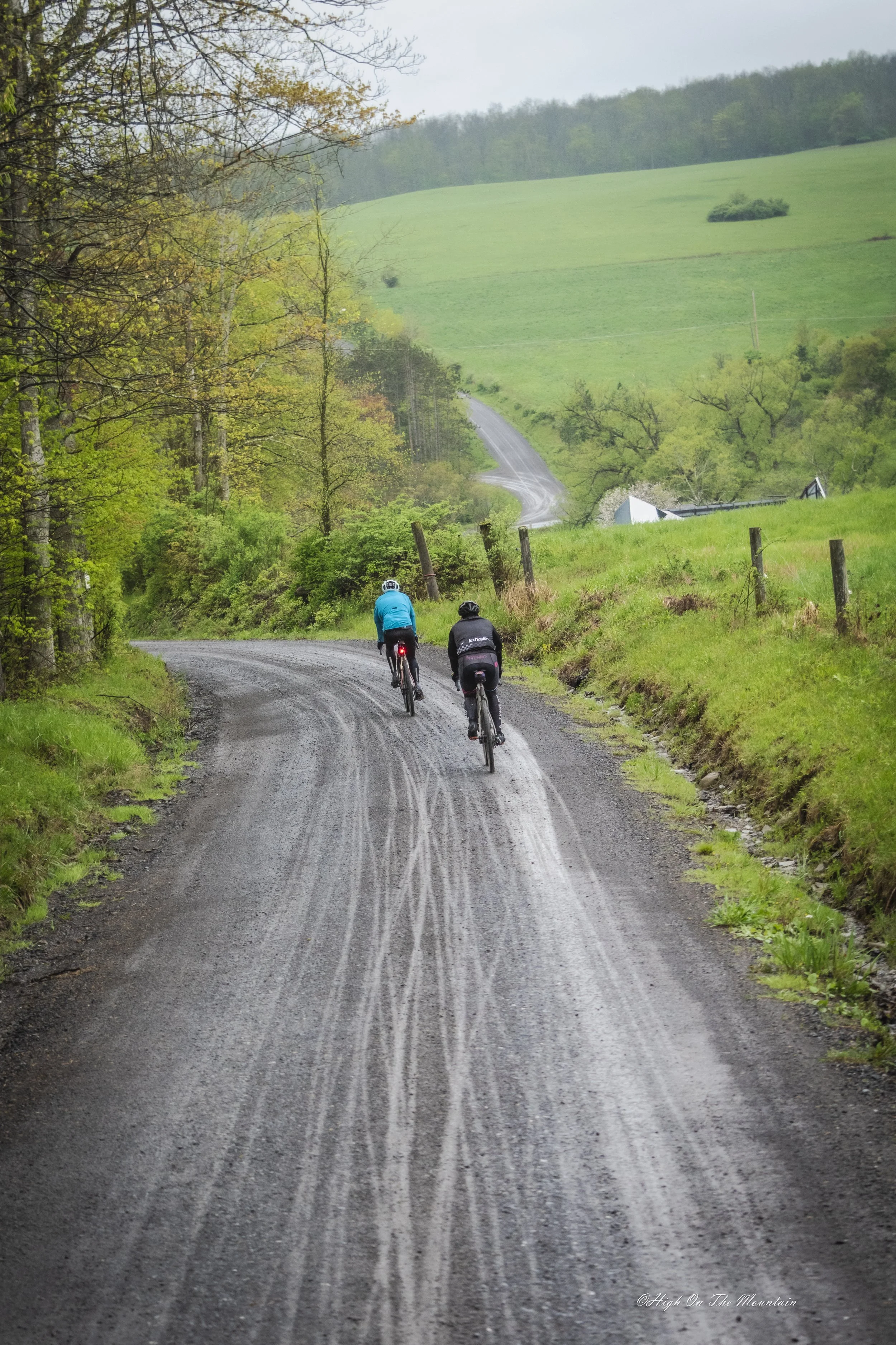 Two cyclists riding on a winding dirt road through a green countryside with trees on the left and rolling hills in the background.
