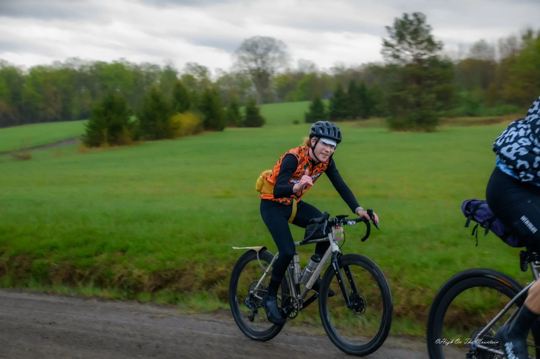 Two cyclists riding on a grassy trail outdoors under cloudy sky, surrounded by trees and greenery.