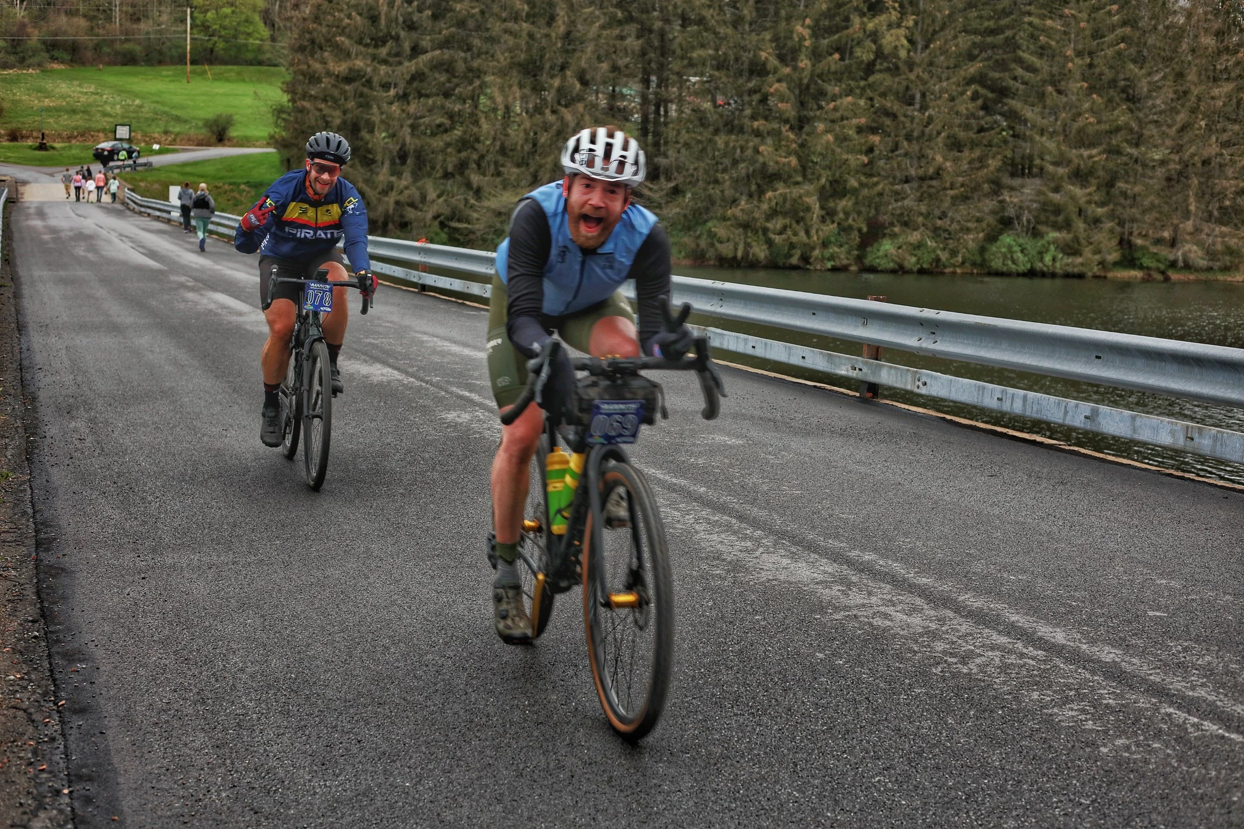 Two men cycling on a paved road beside a body of water with a metal guardrail. The background includes trees and additional cyclists.